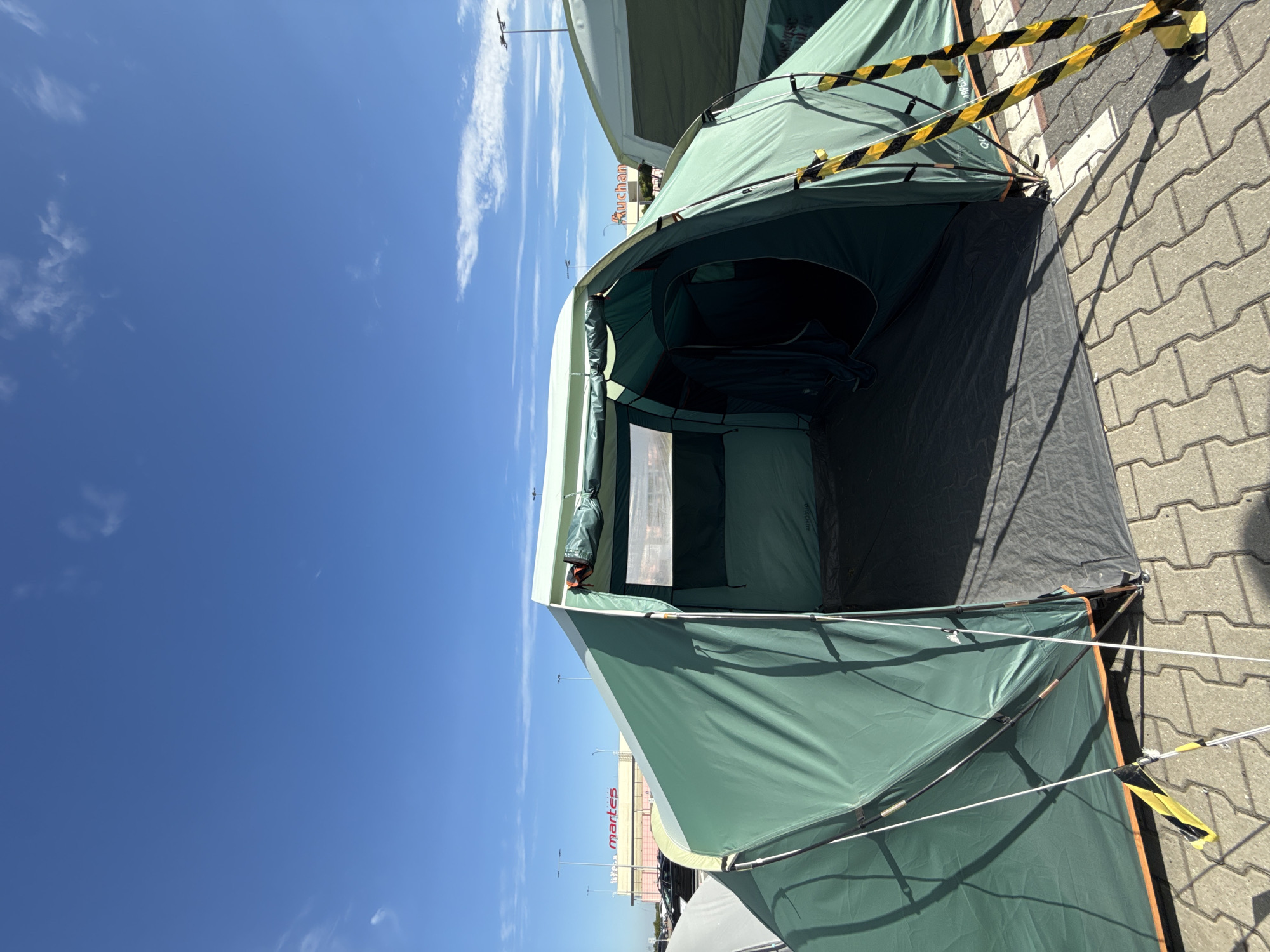 In Bielany Wrocławskie, Poland, a large, dark green tent is set up on a paved area under a clear blue sky with scattered clouds. The tent has an open entrance, revealing a dark interior, and is supported by poles and ropes. Black and yellow caution tape is visible on some of the tent's support structures. In the background, a building with a sign reading "martes" and another with a sign reading "Auchan" are visible, suggesting the location is a retail or commercial area. Several cars are parked in the distance, and streetlights are present. The overall scene depicts an outdoor setting with tents erected, possibly for an event or temporary display, in a retail park environment.