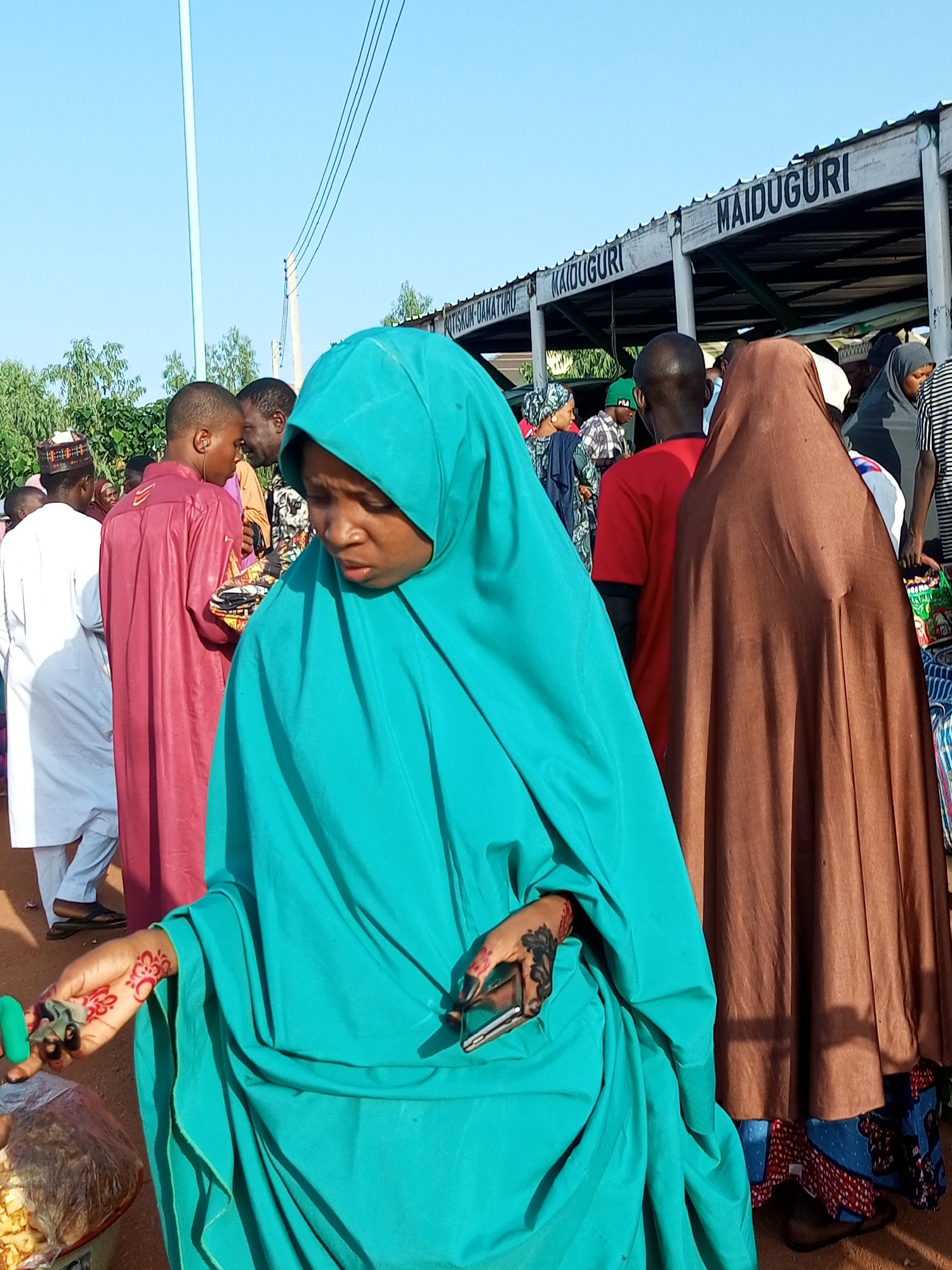 The image depicts a street scene in Gombe, Nigeria, likely at a market or transportation hub. A woman in a turquoise hijab is in the foreground, her hand adorned with red henna. She is holding a small green object and a phone. To her right, a person in a brown hijab stands. In the background, several individuals are visible, some wearing traditional attire such as a maroon kaftan and a white agbada. Overhead, power lines and a structure with signage reading "MAIDUGURI" and "MAIDUGURI DAMATURU" are present, suggesting a connection to travel or commerce. The ground appears to be dirt, and trees are visible in the distance. The overall atmosphere suggests a bustling public space on a sunny day.