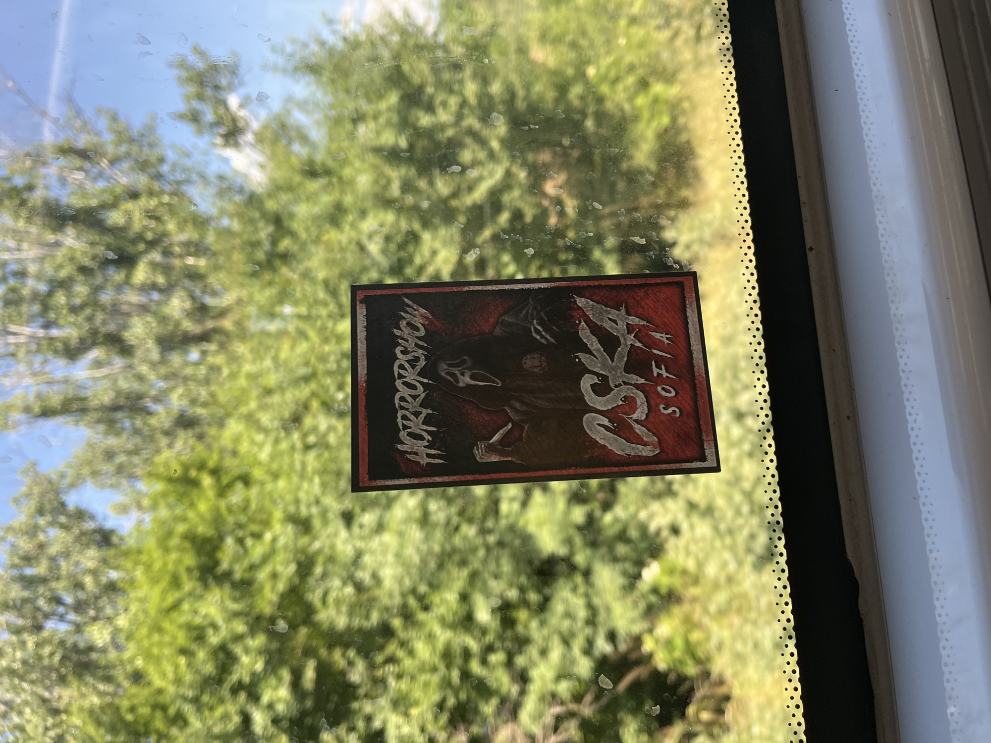 A square sticker featuring the Ghostface mask from the movie "Scream" is affixed to a window, obscuring a portion of the out-of-focus green foliage and blue sky visible through the glass. The sticker has a red border and the text "HORROR SHOW" at the top, with "CSKA SOFIA" in large, stylized white letters at the bottom. The sticker appears to be slightly aged or distressed. The window itself has a dotted line pattern along the bottom edge, and the interior of a vehicle is visible below the window, with a white surface and a hint of grey trim. The setting is outdoors, with lush green trees and a bright sky suggesting a sunny day. This image was captured in Kubratovo, Bulgaria.