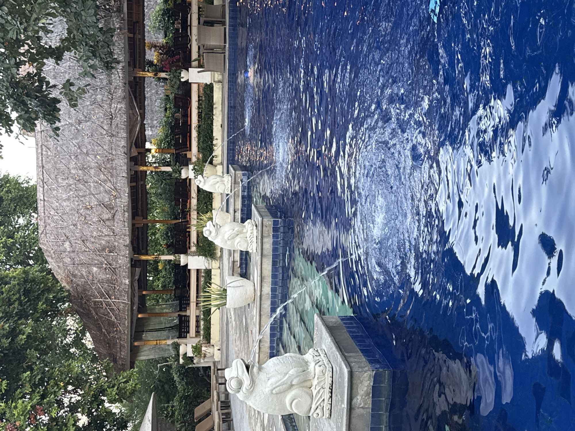 The image shows an outdoor swimming pool with a thatched-roof pavilion in the background. The pavilion appears to be part of a resort or restaurant, with tables and chairs visible. The pool is blue and clean, with several frog-shaped fountains spouting water. One person is swimming in the pool. The location is identified as Denpasar, Indonesia.