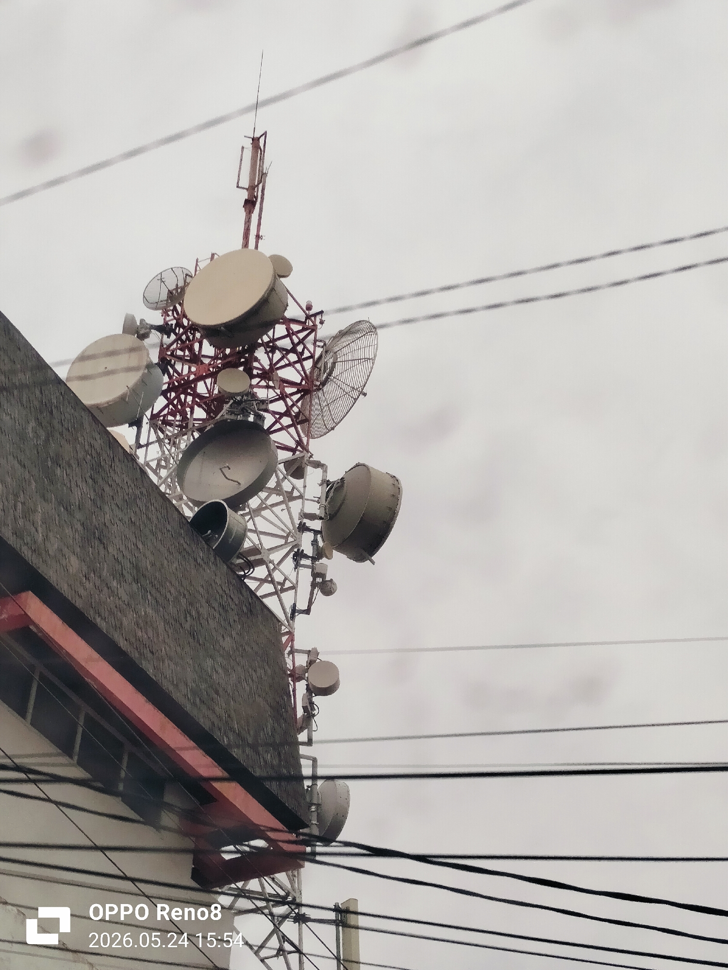 A low-angle view shows a communications tower densely packed with various satellite dishes and antennas, rising from behind a building in Lhokseumawe, Indonesia. The tower's structure is primarily a red and white lattice framework, contrasting with the gray sky and the top of the building, which has a gray brick facade and a red horizontal band. Power lines cut across the foreground, adding to the cluttered composition. The bottom left of the image contains the watermark "OPPO Reno8 2026.05.24 15:54".