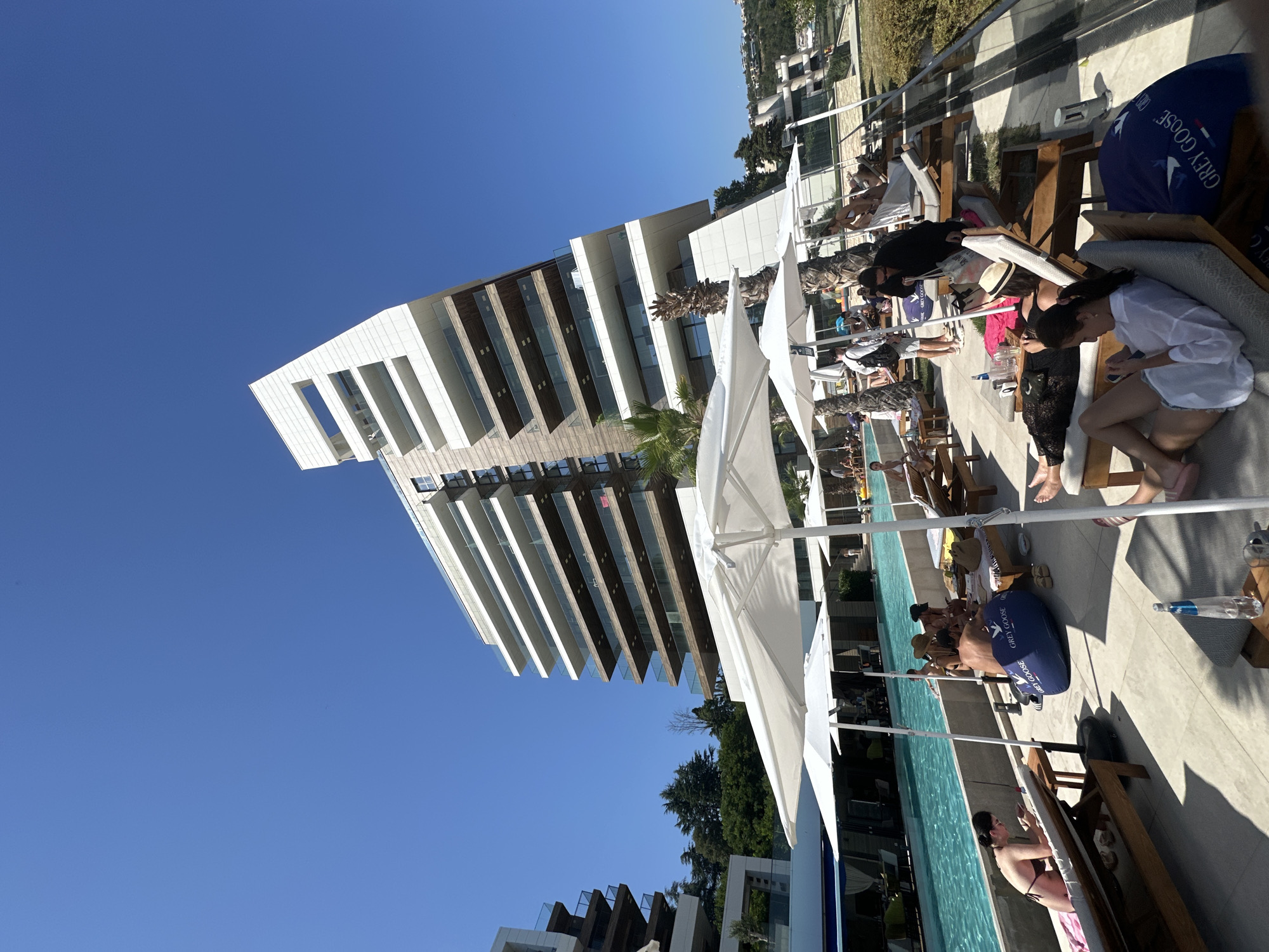 An outdoor shot captures a sun-drenched scene at a resort or hotel. The focal point is a modern, angular building with white and brown facades, its upper floors jutting out over a blue sky. In the foreground, several white umbrellas provide shade for lounge chairs arranged on a tiled patio. A swimming pool with clear blue water is visible to the left, with some people swimming. Various individuals are relaxing on the lounge chairs, some in swimwear, reading, or conversing. One person is lying on a chaise lounge, looking at their phone. Another person is sitting up and appears to be looking at their phone as well. A large blue beanbag chair with a "Grey Goose" logo is placed on the patio. There are several palm trees scattered throughout the scene. The overall atmosphere is one of leisure and relaxation on a bright, sunny day.