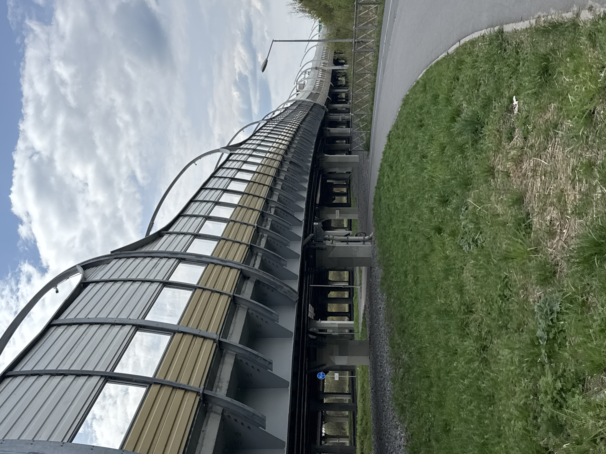 The image shows a curved sound barrier along a road in Saint Petersburg, Russia. The barrier has a metal frame with rectangular panels of reflective material and translucent panels, supported by concrete pillars. The sky is cloudy. Grass and pavement are also visible in the scene.
