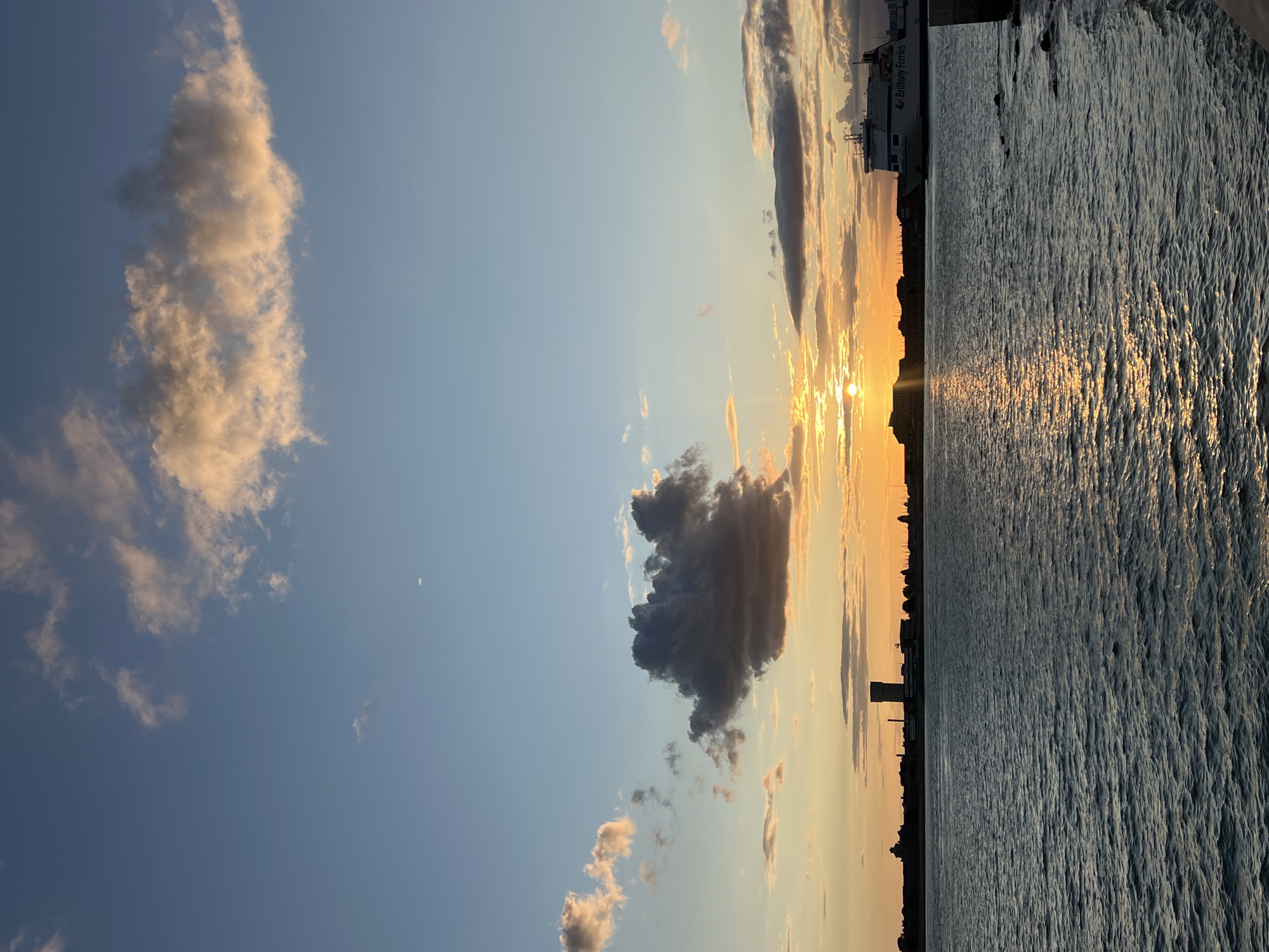A wide shot captures a serene sunset over the waters of Portsmouth, United Kingdom, with the city skyline silhouetted against a vibrant sky. The sun, a bright orb, is partially obscured by the horizon and casts a shimmering golden path across the choppy water. Fluffy clouds, tinged with orange and pink, dot the sky, contrasting with the deep blue above. A large ferry, identified by the "Brittany Ferries" logo, sails serenely in the distance, its white hull stark against the darkening water. The Portsmouth skyline is a dark silhouette, featuring buildings and a prominent tower, likely a landmark. The water is a deep, textured blue, reflecting the warm hues of the sunset and the sky. The scene is tranquil, evoking the end of a day in the historic maritime city.