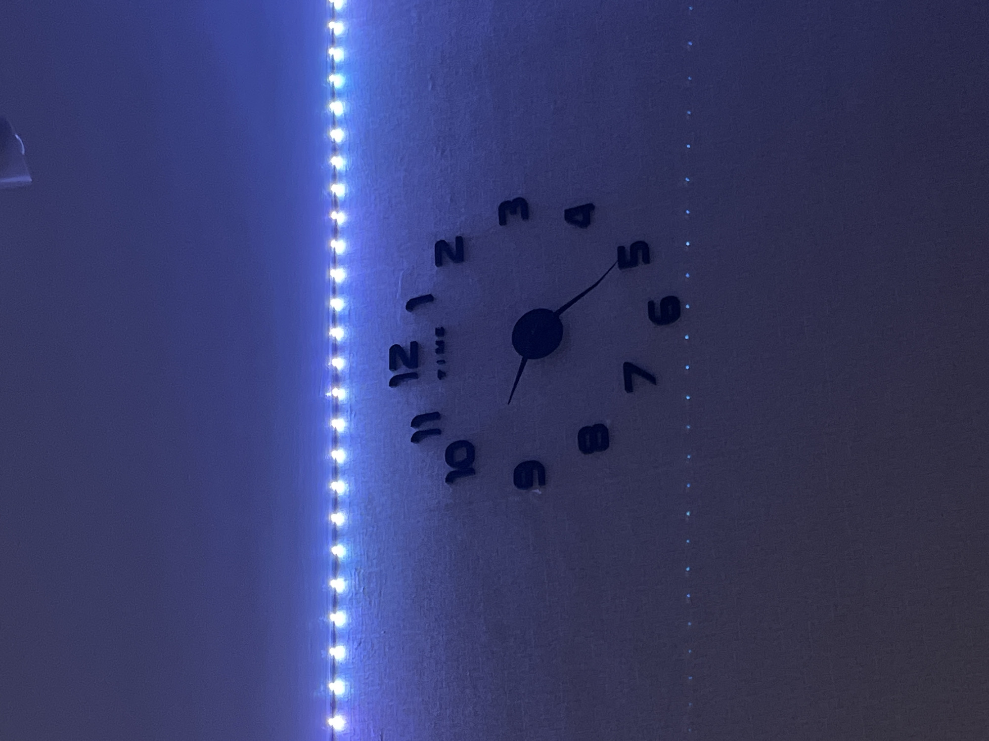 The image depicts a wall clock with black numerals and hands indicating a time around 5:30. The wall is lit with blue LED strip lights on the left and what appears to be a strand of small lights on the right. The environment appears to be a dimly lit interior space. The location in Chaikovsky, Russia is not discernible from the image.