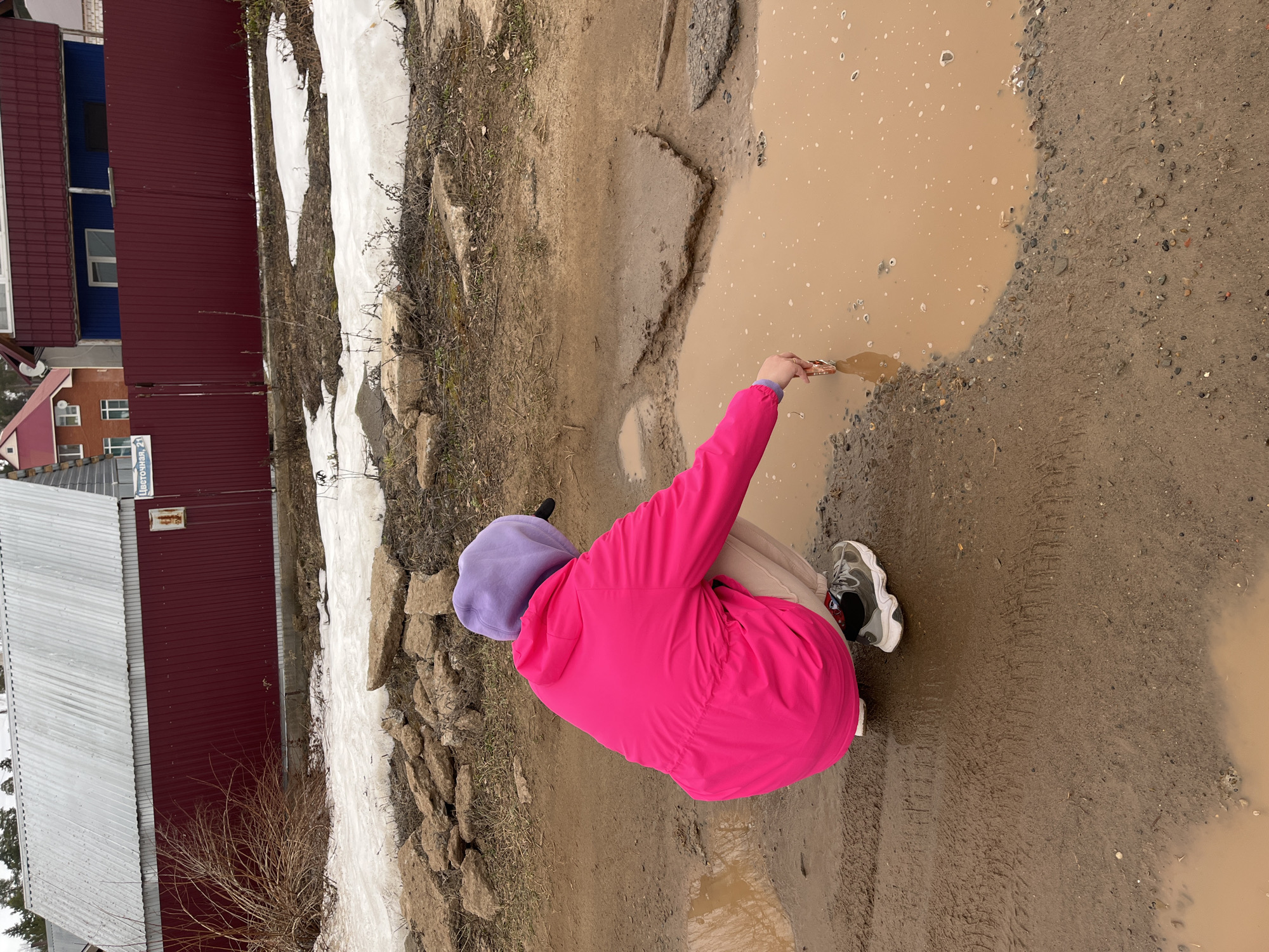 A person in a pink jacket and purple hood squats next to a large, muddy puddle. The person's right hand is reaching into the puddle. The ground around the puddle is muddy and has tire tracks. Patches of melting snow are visible near a red and grey building in the background. The building has a sign on the front that is partially obscured but appears to read "цветоч." This suggests the location is likely near a flower shop. Based on the partial sign and the prompt, the location is likely in the city of Chaikovsky, Russia.