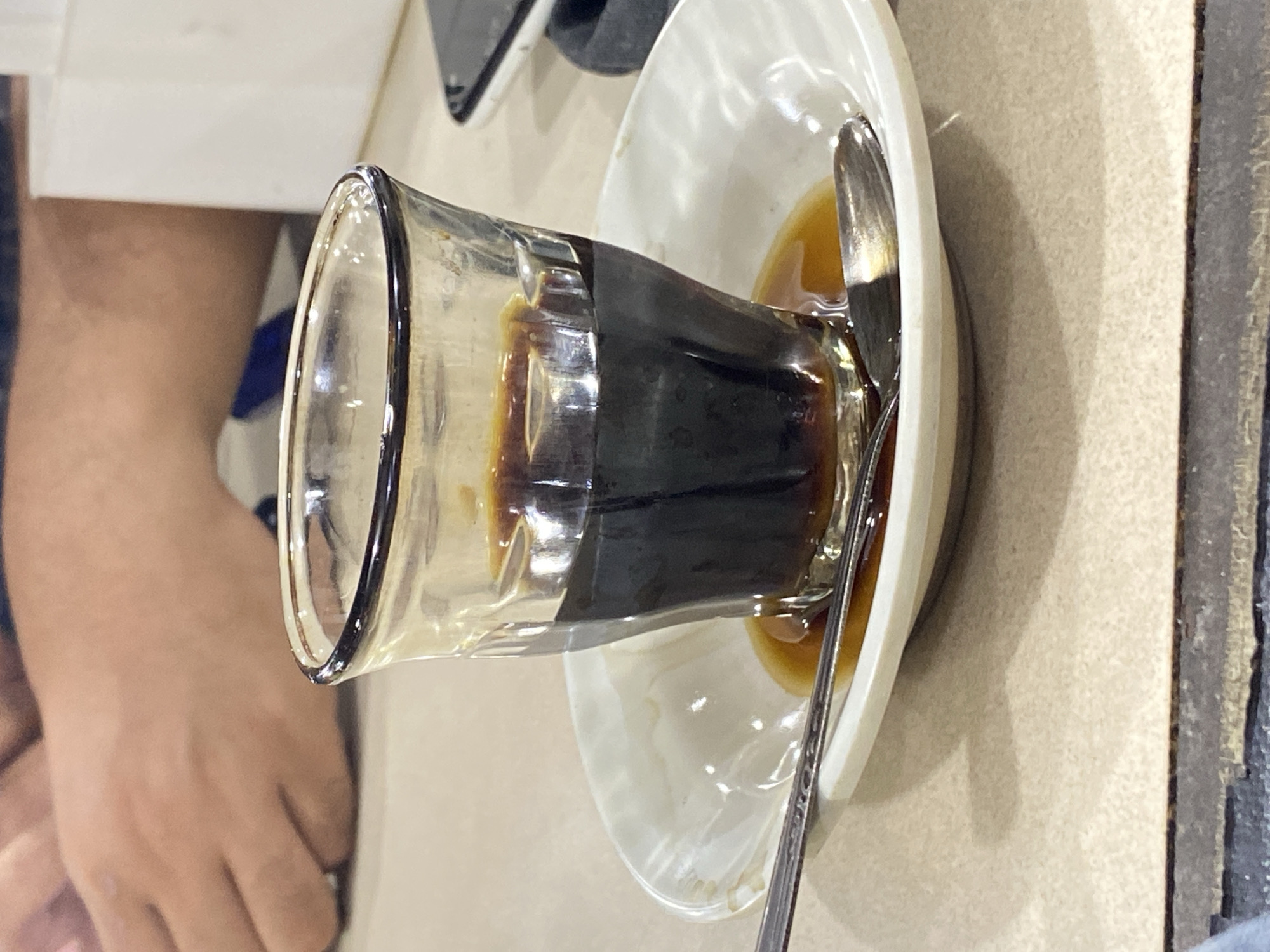 The image shows a close-up of a small glass filled with dark liquid, likely coffee or tea, placed on a white saucer. A silver spoon rests on the saucer, partially submerged in the liquid. The glass has a thick rim and is slightly tilted, with some of the liquid spilled around the base on the saucer. In the background, a person's arm and hand are visible, suggesting they are holding or about to pick up the glass. The setting appears to be an indoor cafe or restaurant, with a textured beige surface visible. The lighting is dim, creating a warm and inviting atmosphere, possibly indicating an evening or early morning setting. The overall mood is relaxed and suggests a moment of quiet enjoyment of a beverage. There is no visible text in the image. The crops provide a closer look at the glass rim, the liquid, the spoon, and the person's hand. The location context suggests this is a scene from Lhokseumawe, Indonesia, which could imply a specific type of local beverage or serving style.