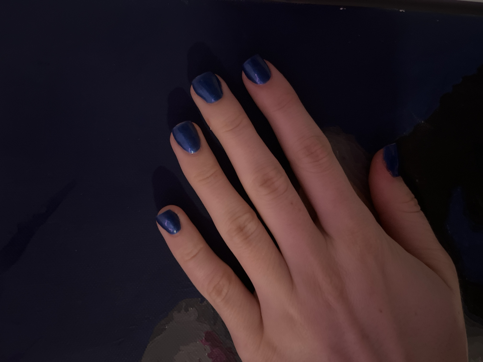 A fair-skinned human hand is positioned on a dark blue, somewhat reflective surface. The hand features fingernails painted with a matching dark blue, possibly metallic, nail polish. The thumb, index, middle, ring, and pinky fingers are visible. The background is out of focus. The location of the image is not discernable from the image.