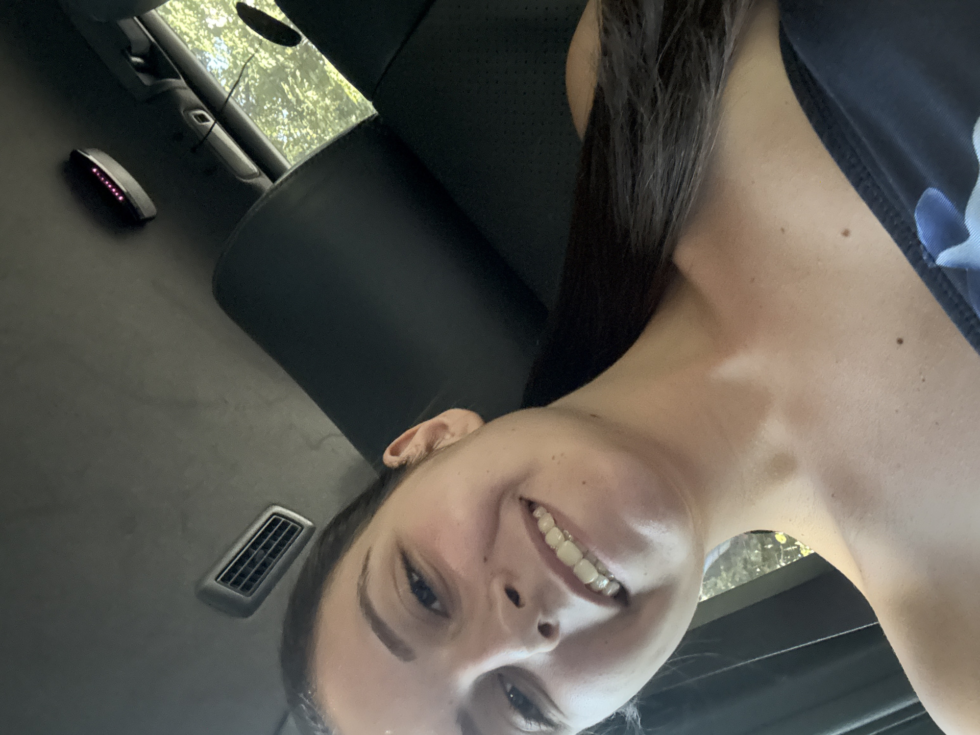 This media file is a close-up selfie taken inside a car. The primary subject is a young woman with long, dark hair, wearing a dark top with a floral pattern. She is smiling at the camera, with her head tilted.
In the background, the interior of the car is visible. This includes a black leather seat, a car vent, and a small, oval-shaped object with several pink lights. Above and behind her, a sunroof reveals a view of green foliage, suggesting the car is outdoors in a leafy area. The lighting suggests it is daytime.
The image is shot from a low angle, looking up at the woman. The overall mood is casual and happy, indicated by her smile. There is no visible text in the image. The location context provided is Burgas, Bulgaria, but this cannot be visually confirmed from the image itself.