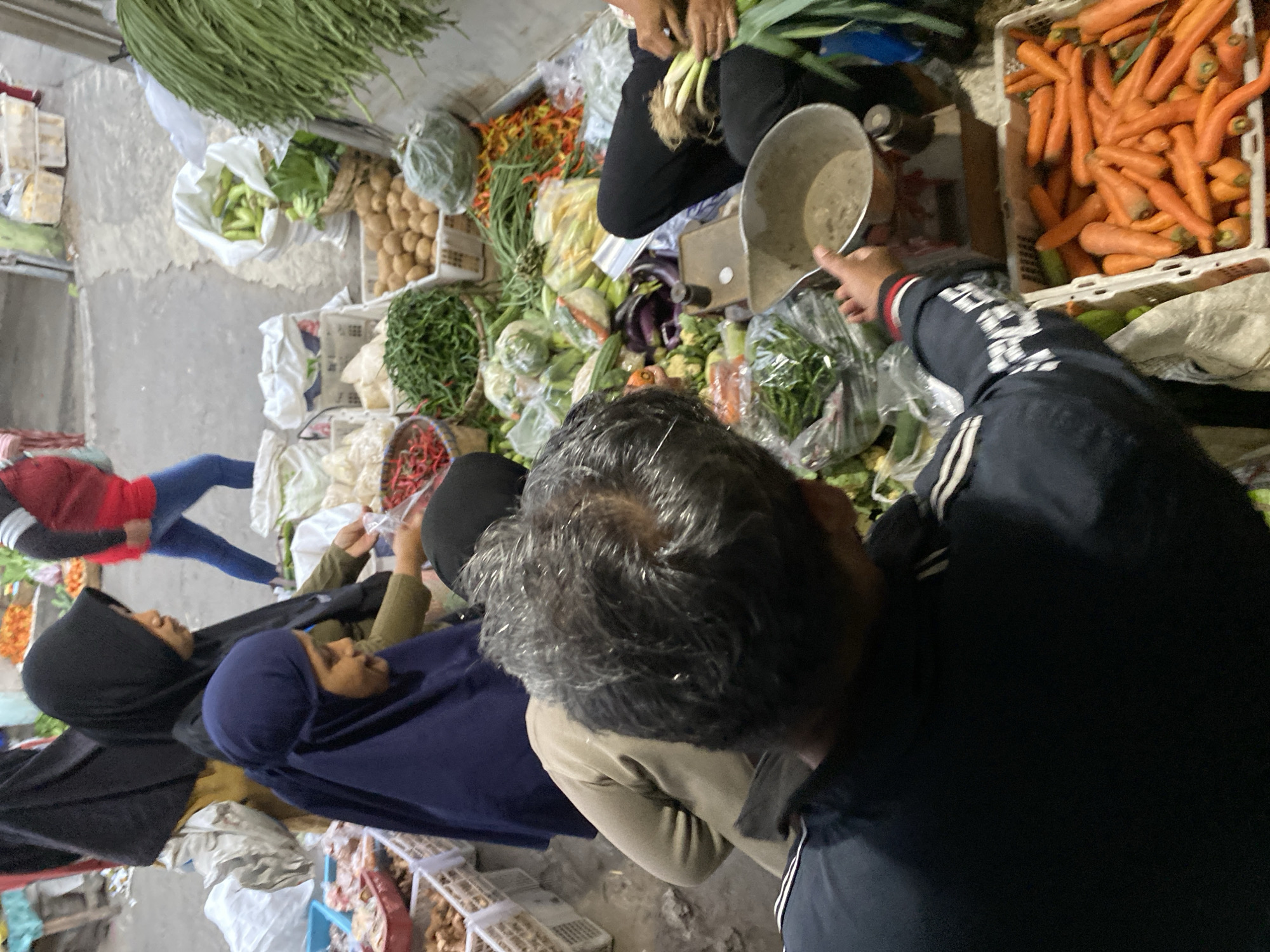 A group of people are gathered around a market stall in Ambarketawang, Indonesia, engaged in shopping. The stall is laden with a variety of fresh produce, including carrots, chilies, long beans, and other vegetables. In the foreground, a man wearing a dark blue jacket with white stripes on the sleeves is seen from behind, interacting with the stall. To his left, a woman in a blue hijab is looking down. Behind her, another woman in a black hijab is also present. In the background, a person in a red shirt and blue pants is visible, along with various market stalls and goods. The ground appears to be paved, suggesting an outdoor market setting.