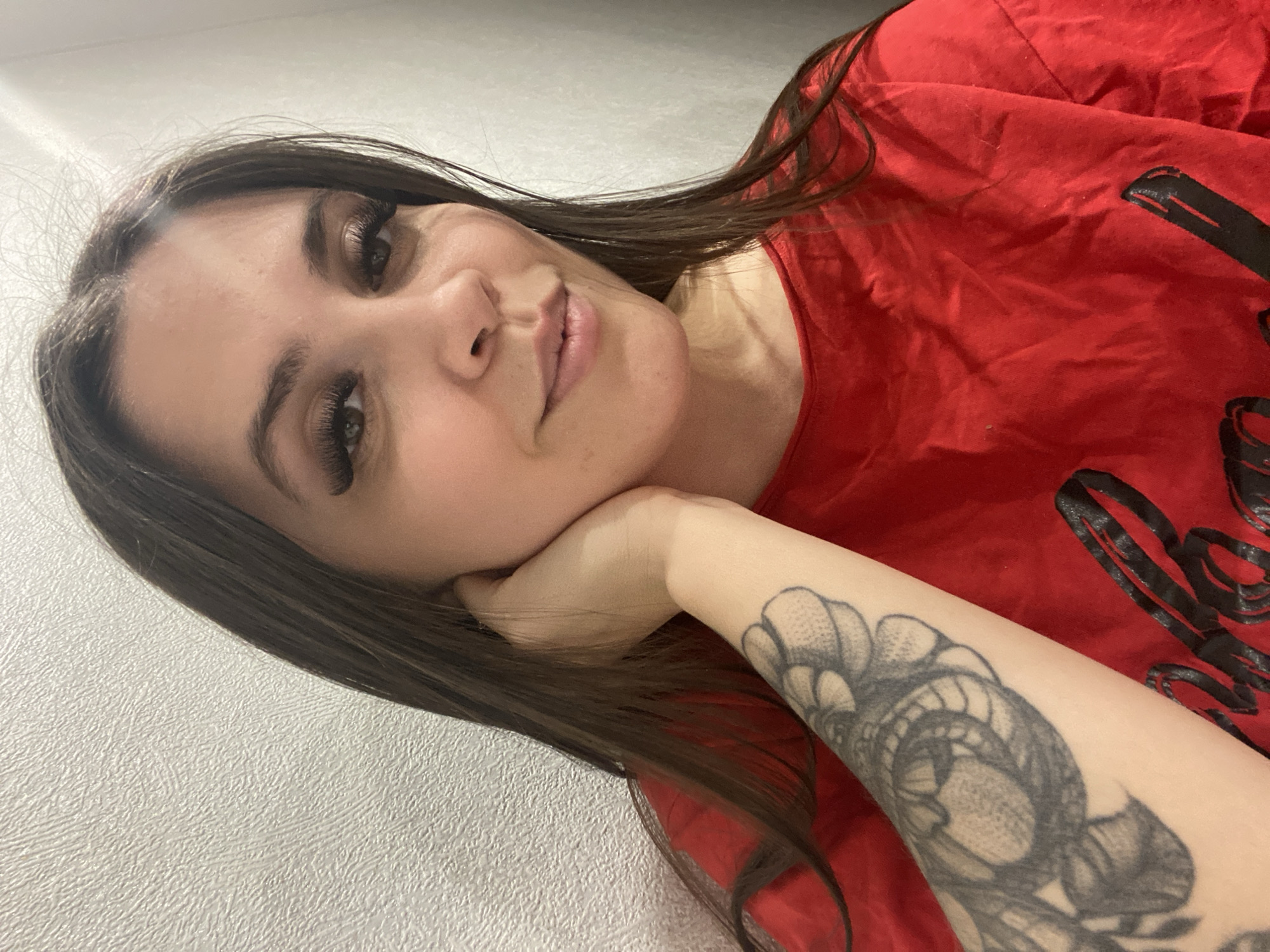 A woman with long brown hair and extended eyelashes is lying down, resting her face on her left hand, with a large, gray-scale floral tattoo visible on her forearm. She is wearing a red t-shirt with a large, black graphic design printed on it. The background consists of a plain, light-colored wall. The location is not discernible from the image.