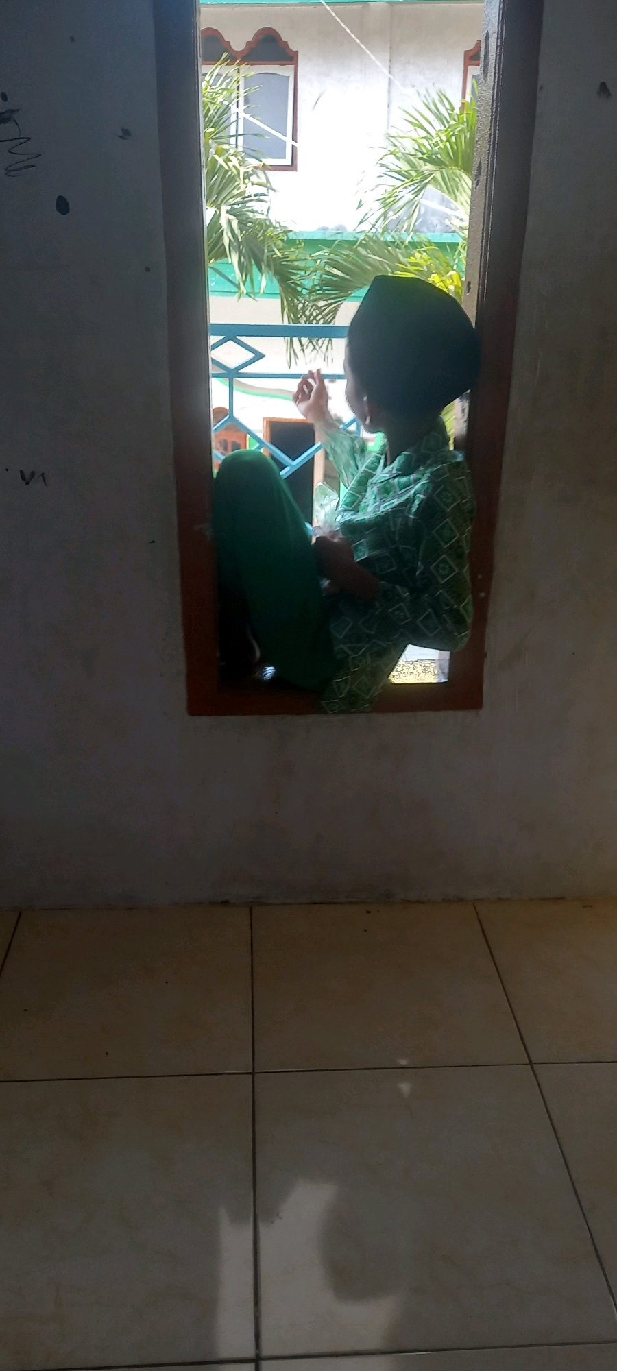 A child wearing a green patterned shirt and green pants sits on a windowsill looking out. The child is wearing a black head covering. The window is open and shows palm trees and a building in the background. The room is sparsely furnished with a tiled floor and a rough-textured wall. The image appears to have been taken in Pontang, Indonesia.