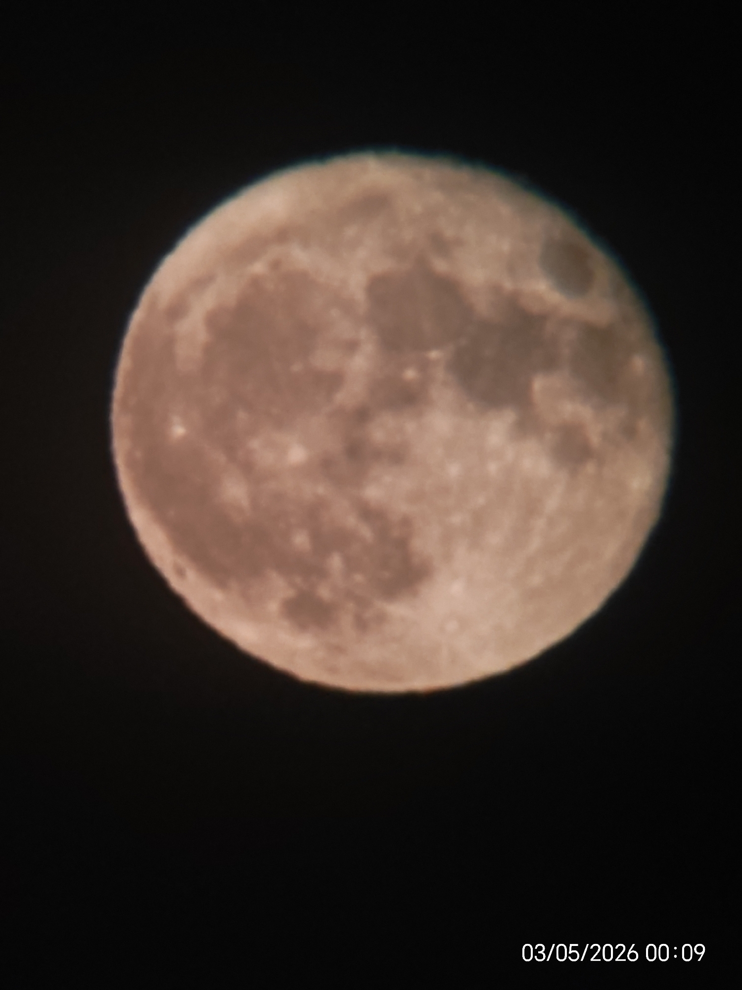 A close-up photograph shows the moon against a black background. The moon is nearly full and its surface is visible with craters and varying shades of grey and pink. The date "03/05/2026 00:09" is printed at the bottom right of the image. The location is unknown.