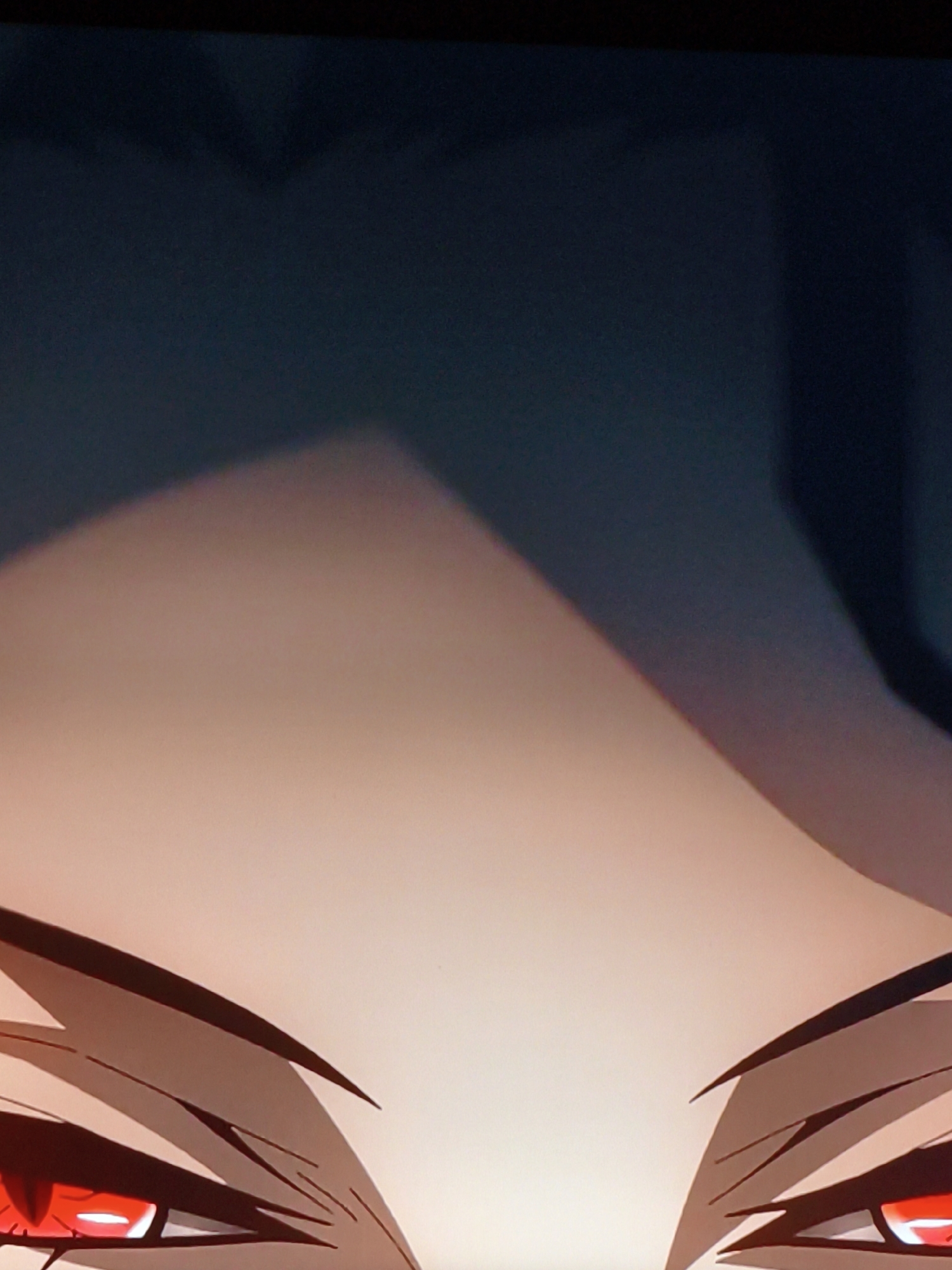 The image is a close-up, low-angle shot of an anime-style character's face. The character has intensely red eyes with bright white highlights, suggesting a glow or supernatural power. Their eyebrows are sharply angled and dark, and there are visible dark lines that appear to be eyelashes or stylized facial features. The skin tone is a pale peach. Above the eyes, a shadow creates a triangular shape that falls across the forehead, implying the presence of hair or a headdress casting the shadow. The background transitions from the character's skin tone to a dark, shadowy blue-grey, obscuring any details about the environment or location. There is no discernible context to place this image within Vladivostok, Russia.
