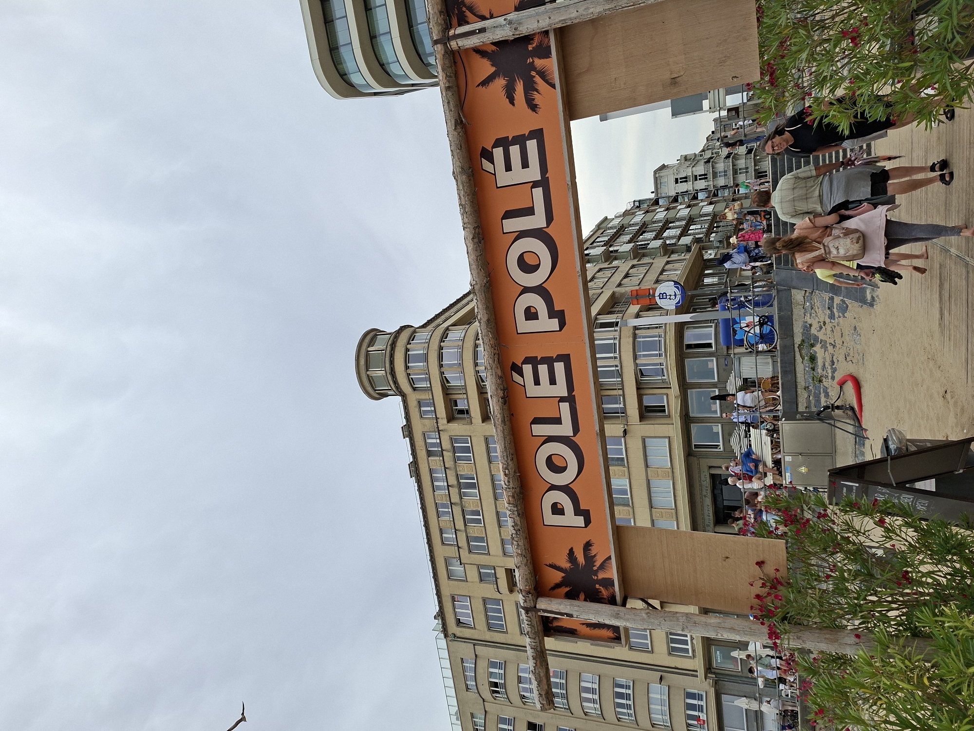 An outdoor, eye-level, medium shot captures the "POLE POLE" sign, a beach entrance, and buildings along the Ostend, Belgium coast on a cloudy day. The sign, set on wooden posts, features orange lettering with white outlines, accented by silhouetted palm trees. Behind the sign, an apartment building with many windows stretches across the background, its tan facade contrasting with a modern, curved white building on the right.
In the foreground, a sandy path leads through lush green plants with red flowers. A few people are visible walking on the path towards the beach entrance. Further back, more people are gathered near the building's entrance, some sitting on a bench under striped umbrellas. A seagull is captured in flight in the upper left corner of the frame. The overall scene depicts a lively beachside atmosphere.