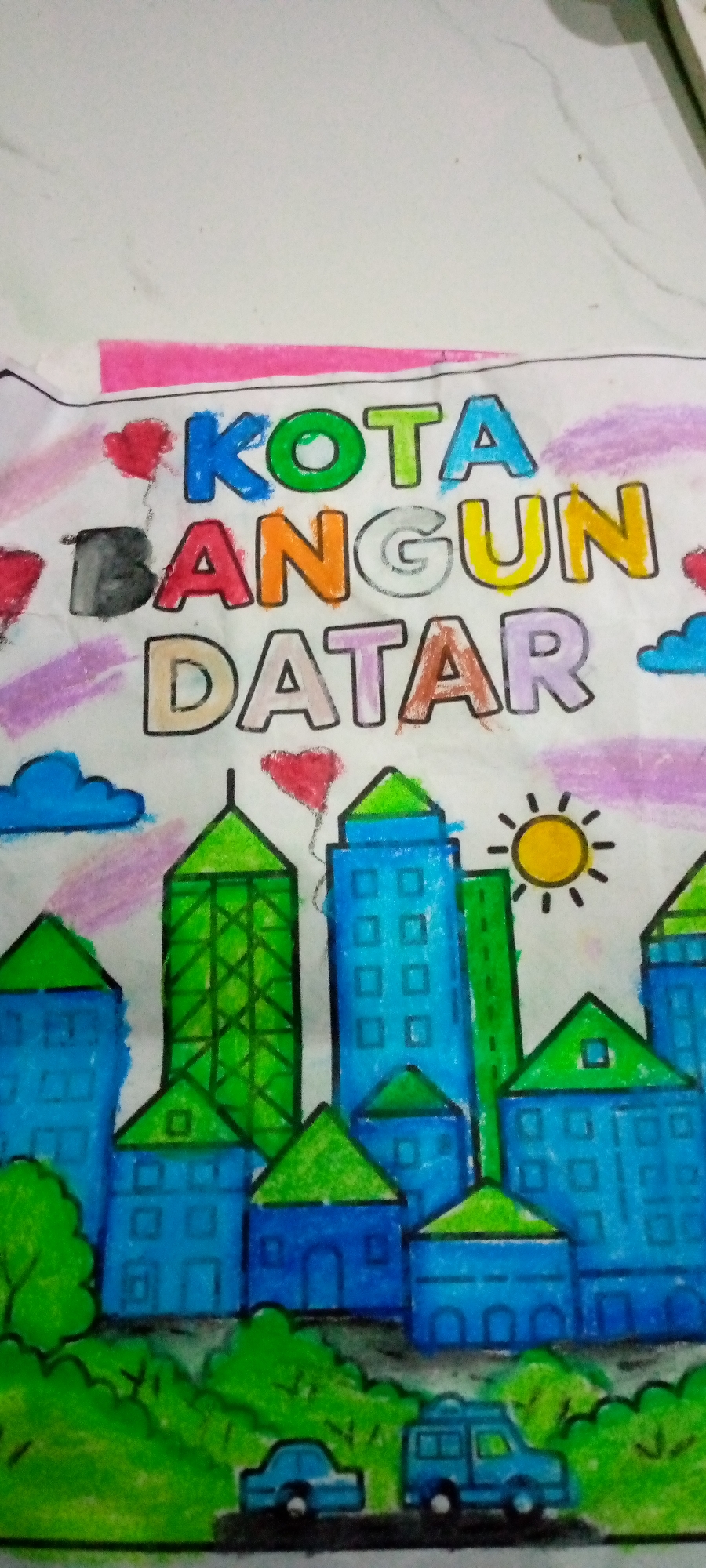 The image is a colorful drawing of a city skyline. The text "KOTA BANGUN DATAR" is prominently displayed across the top, written in large, block letters of various bright colors. Below the text, a cityscape unfolds with stylized buildings. These buildings are depicted in shades of blue and green, with some featuring green triangular roofs. There are also elements like clouds, hearts, a sun, and trees scattered throughout the drawing. In the foreground, two simplified cars are shown on what appears to be a road or ground. The overall style is childlike and appears to be drawn with crayons or colored pencils on a white surface. There is no specific indication of the location being Sumberwono, Indonesia within the visual content of the drawing itself.