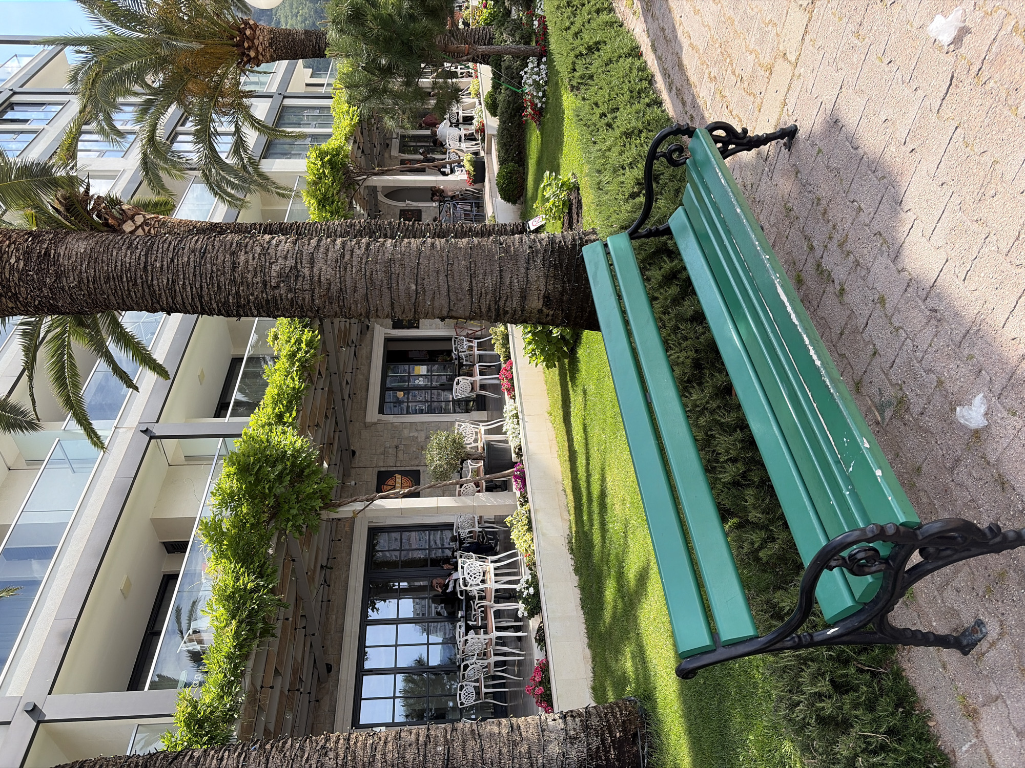 The scene is set in Rafailovići, Montenegro. A green wooden park bench with black iron legs sits on a brick walkway, adjacent to a grassy area. A large palm tree is positioned near the bench. In the background is a building with glass-paneled balconies and a restaurant with outdoor seating. People are seated at the restaurant tables. Flowers and greenery surround the outdoor seating area. The image is taken at a slightly oblique angle with the camera tilted to the side.