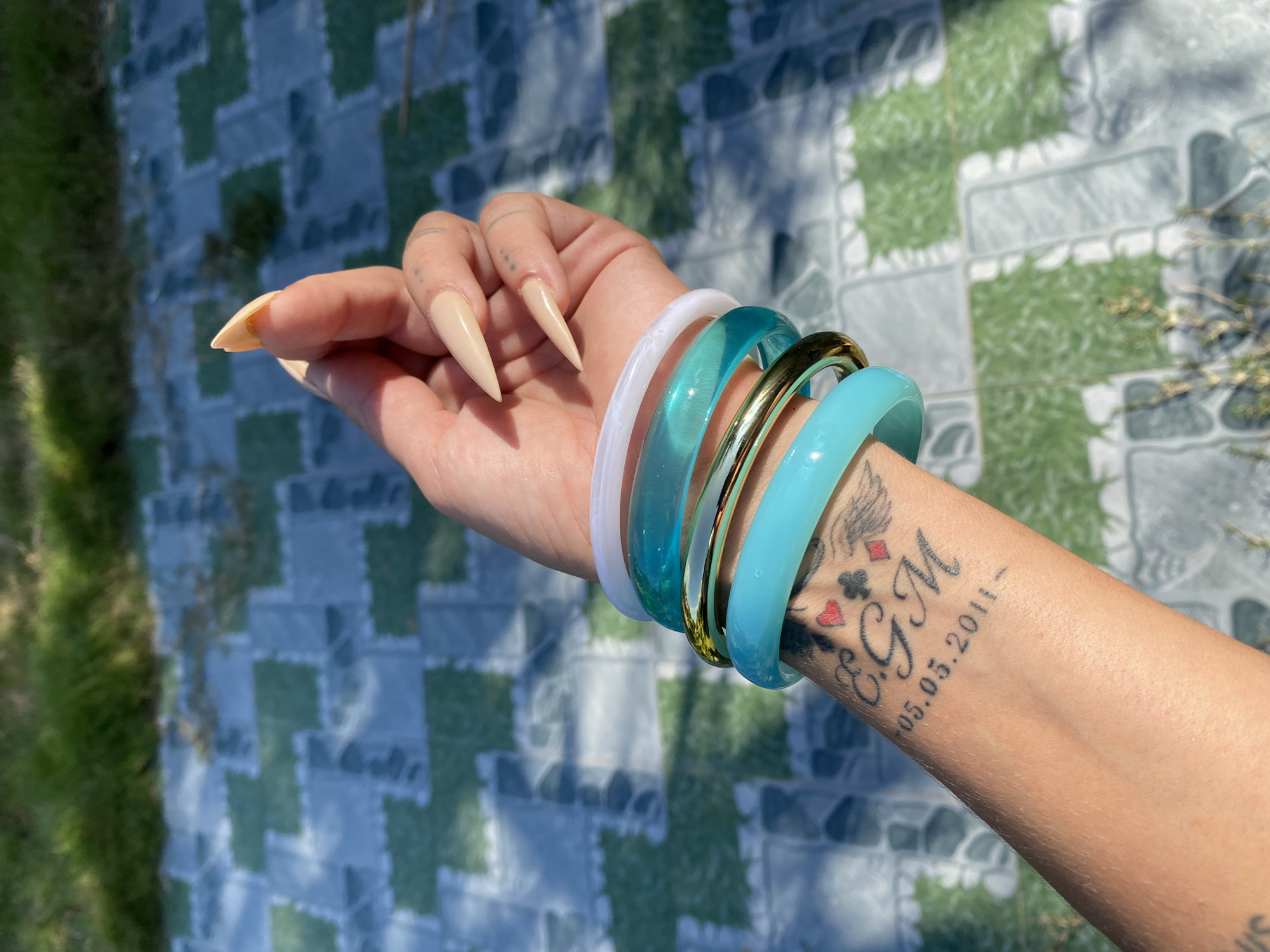 A person's forearm and wrist are adorned with several translucent bangles of varying shades of turquoise and white, along with one gold bangle. The bangles are stacked on the wrist, covering a tattoo that appears to depict playing card suits and the initials "EGM" with a date "05.05.2011" below it. The person's hand, with long, pointed nails painted in a nude color, is visible in the upper right corner, held near the bangles. The background consists of a tiled surface with a repeating pattern of green and grey squares, suggesting an outdoor setting. Above the tiled area, green grass is visible, illuminated by sunlight that casts shadows on the ground. This scene could be located in the city of Mũi Né, Vietnam, potentially at a resort or outdoor seating area, given the combination of tiled patios and natural greenery.
