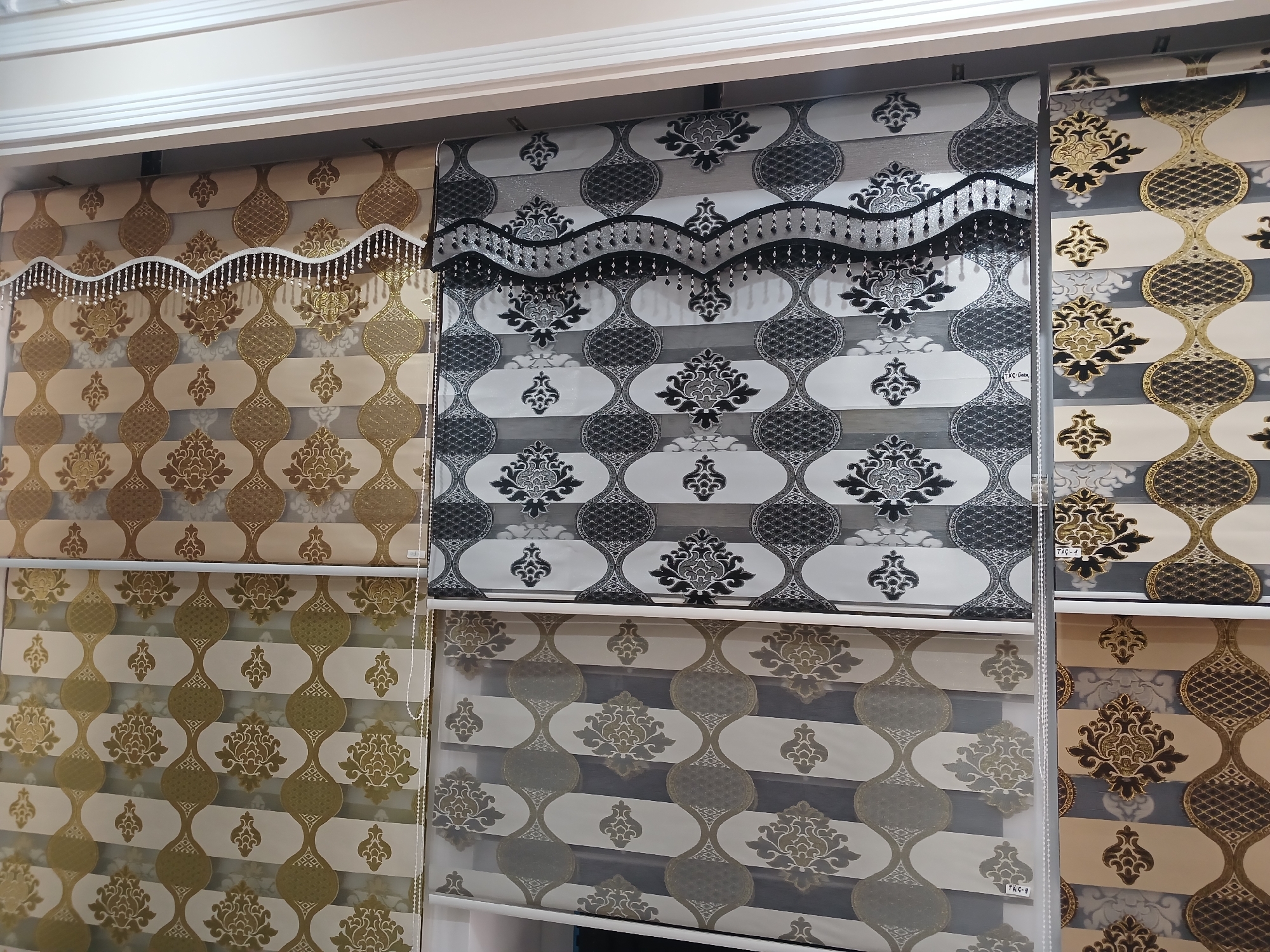The image shows a retail display of roller blinds in a store located in the city of Türkmenabat, Turkmenistan. The blinds feature intricate damask patterns in gold and beige, and black and white color schemes. Some blinds have decorative beaded trim along the top. The blinds are displayed in vertical rows, with each row showcasing a different pattern and color combination. The overall environment appears to be a clean and well-lit showroom.