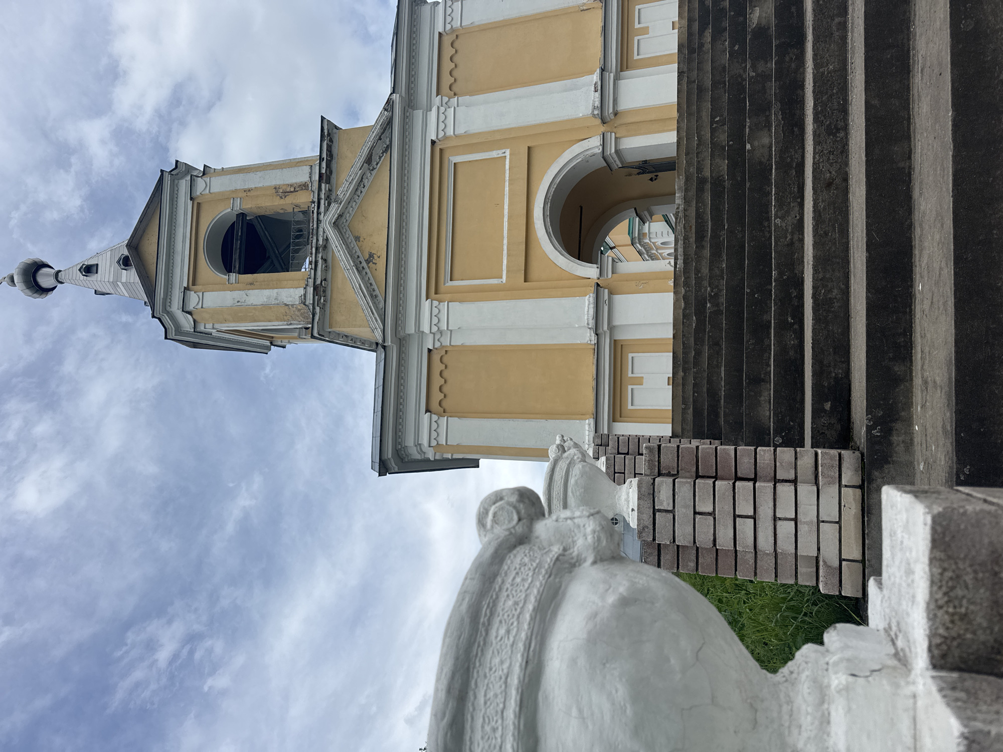 The image shows a low-angle view of a yellow bell tower with white trim, against a cloudy sky. In the foreground, there are concrete stairs leading up to an arched entryway of the bell tower. A large white urn and a brick pillar are visible on the left side of the stairs. The bell tower appears to be part of a religious building, possibly a church or monastery, given its architectural style and the presence of a dome at the very top of the tower. The overall scene suggests an outdoor location, likely in the city of Рассвет, Russia, as indicated. There are no people visible in the image.