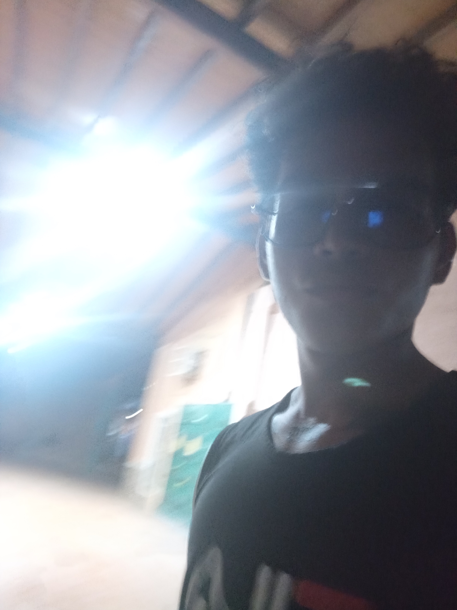 The image is a selfie taken by a person wearing dark sunglasses and a black t-shirt. The background appears to be an indoor space with wooden beams overhead. There is a bright light source to the left of the frame, creating a glare. The person's face is partially obscured by the sunglasses and the lighting. The lower portion of the image shows the person's chest and t-shirt. The scene does not provide any specific visual cues to indicate its location in Juan Griego, Venezuela.