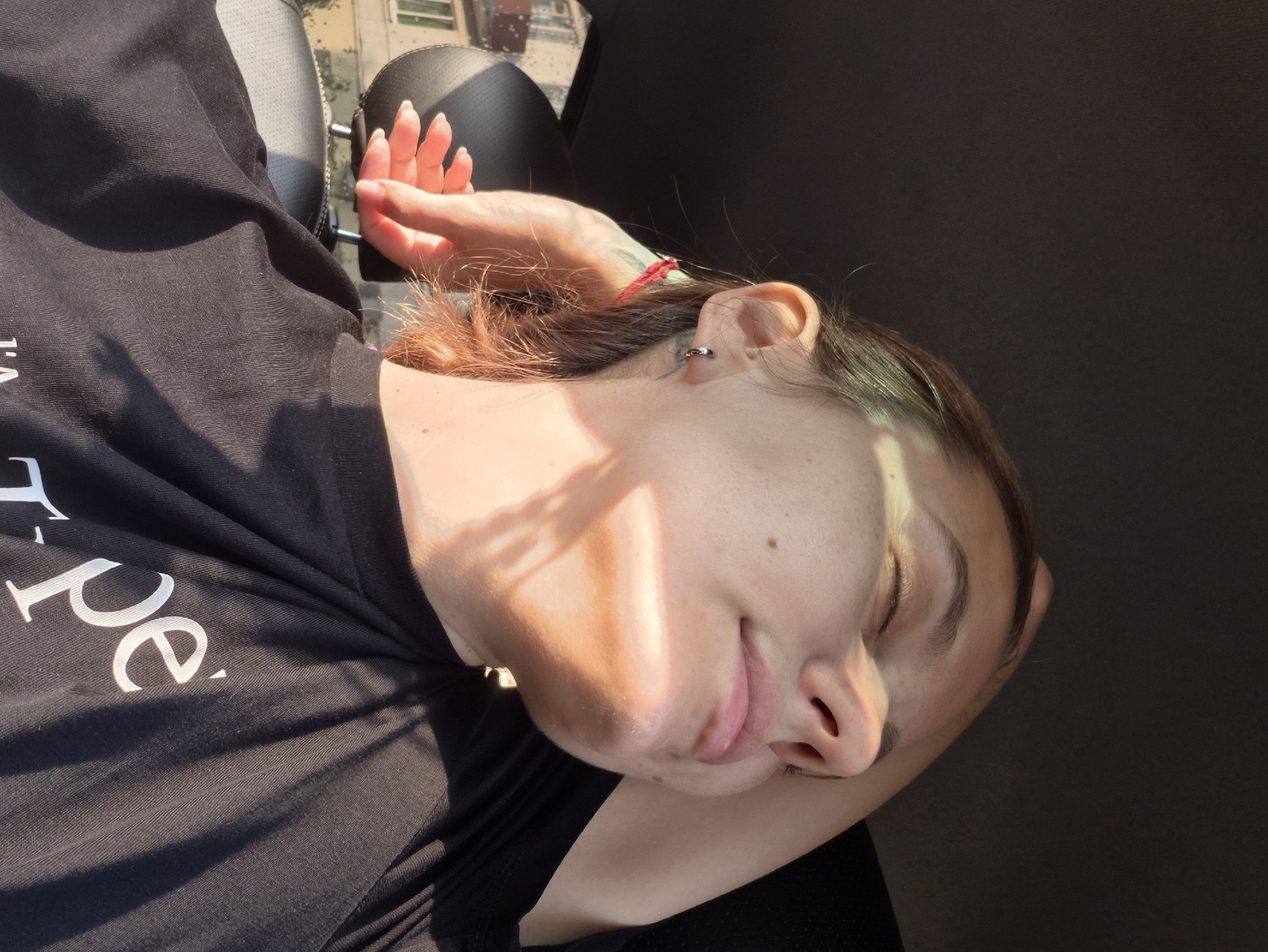 A young woman is sitting in the passenger seat of a car, facing the right side of the frame. She has her eyes closed and her right arm is raised, with her hand resting on her head. Sunlight is casting shadows across her face. She is wearing a black t-shirt. The interior of the car is visible, including a headrest and a portion of the ceiling. Outside the car window, buildings and trees can be seen. The image appears to have been taken in the city of Chaikovsky, Russia.