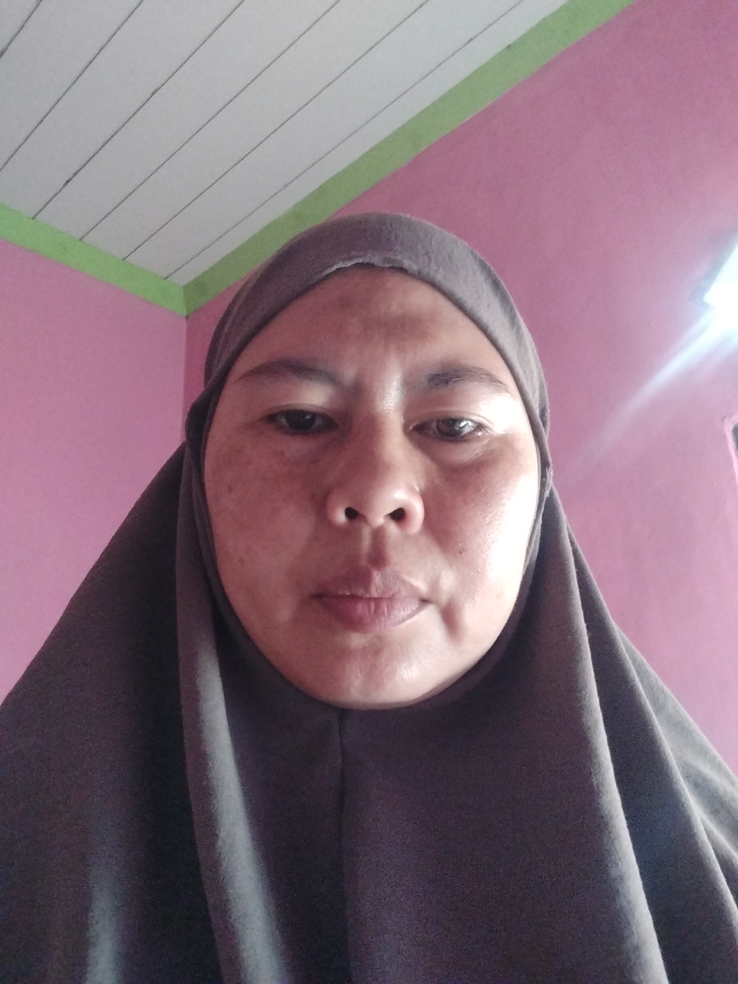 An over-the-shoulder, head-on selfie is taken from a low angle, looking up at the face of a woman wearing a dark gray hijab. The woman's face is centrally framed, with her head tilted slightly forward. Her skin appears to have freckles and some texture. The background consists of a pink wall and a ceiling with white wooden panels, separated by a light green trim. The lighting appears to be from a bright source to the right of the frame, casting a glare on the wall and the woman's face. The scene suggests an indoor domestic setting, likely a room within a house in Daya Asri, Indonesia, given the style of the ceiling and the typical colors used in residential interiors in the region.