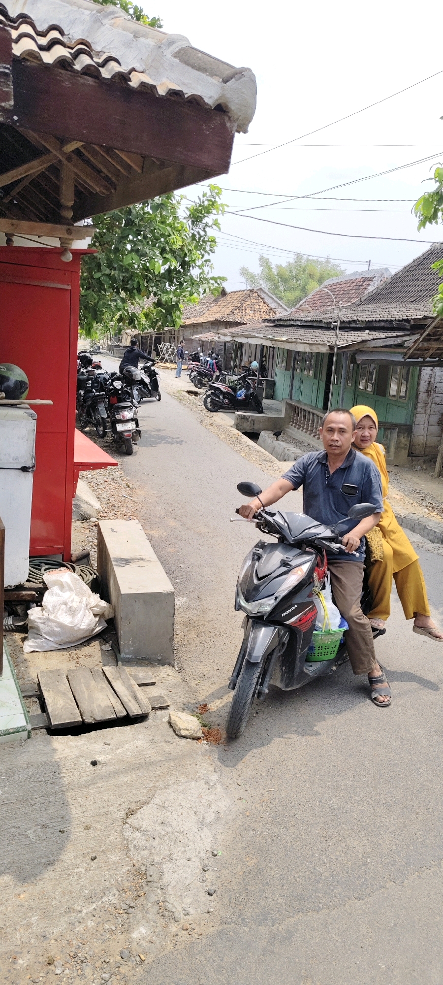 This is a medium shot, taken from a low angle, of two people riding a motorcycle on a street in Lambangan Kulon, Indonesia. A man is driving the motorcycle, and a woman is sitting behind him. The man is wearing a dark blue polo shirt and brown pants. The woman is wearing a yellow hijab and yellow pants. They are both looking at the camera.
The motorcycle is black and has a green basket attached to the front, containing a white plastic bag and some other items. The motorcycle has the brand "Honda" on its front. The man is wearing dark sandals, and the woman is wearing light-colored sandals.
The street is paved and appears to be in a rural or suburban area. On the left side of the street, there is a red building with a wooden roof overhang. Next to the building, there is a concrete block, a pile of white sacks, and some wooden planks laid over a hole in the ground. On the right side of the street, there are several small buildings with wooden exteriors and tiled roofs, characteristic of the architecture in the region. A row of parked motorcycles is visible further down the street. Trees and overhead wires are also present in the background. The sky is overcast.