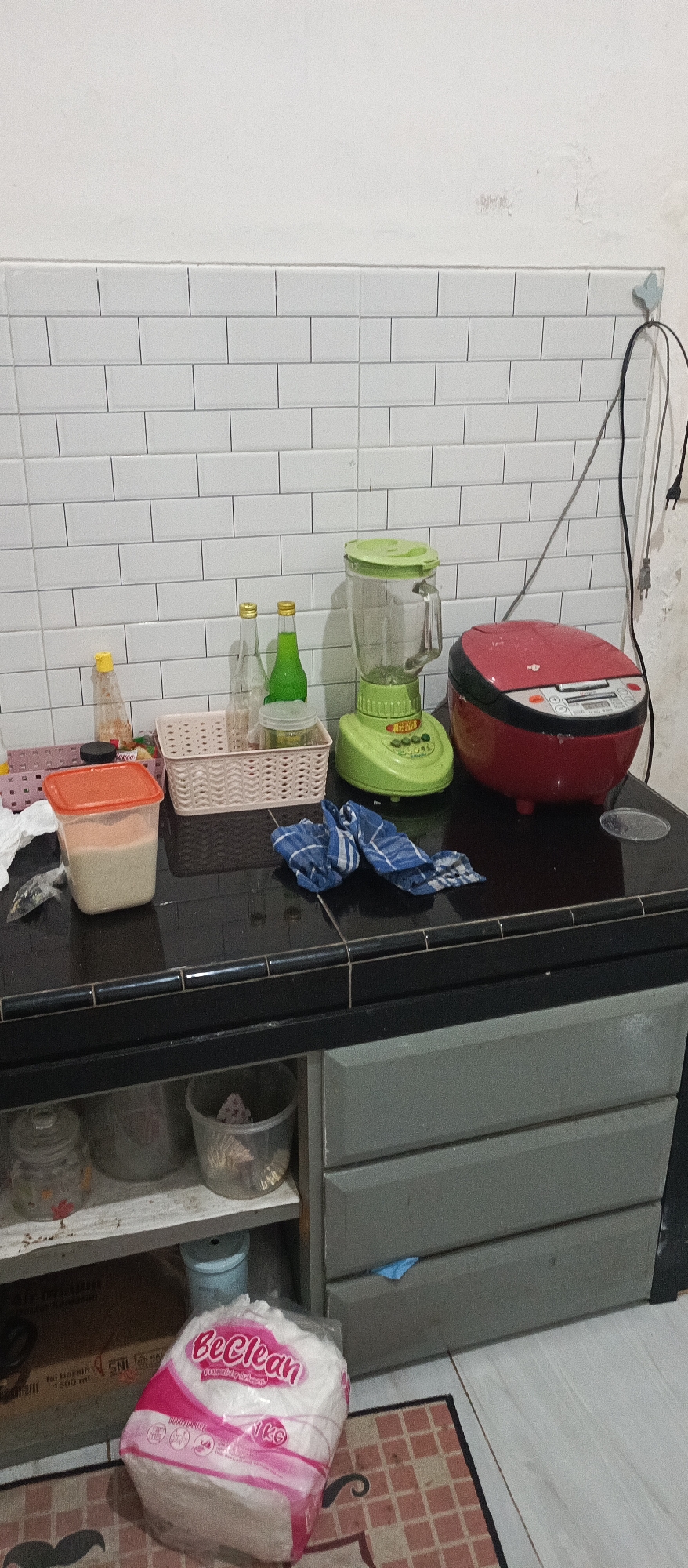 In a kitchen area that appears to be in Arjasa, Indonesia, a countertop is set with various kitchen appliances and containers. A red rice cooker sits to the right of a green blender. In front of the blender, there are two glass bottles, one containing a clear liquid and the other a green liquid. To the left of the bottles is a small, clear jar. Next to this, a woven basket holds an orange-lidded plastic container filled with a light-colored powder. A blue and white checkered cloth is placed on the countertop near the blender. Below the countertop, a gray cabinet with three drawers is visible. On a shelf beneath the countertop, a large glass jar and a clear plastic container holding what appear to be cupcake liners are stored. To the left of these items, a cardboard box is partially visible. In the foreground, a large, white bag of tissues with pink lettering that reads "BeeClean" is placed on the floor. The wall behind the countertop is tiled with white subway tiles.