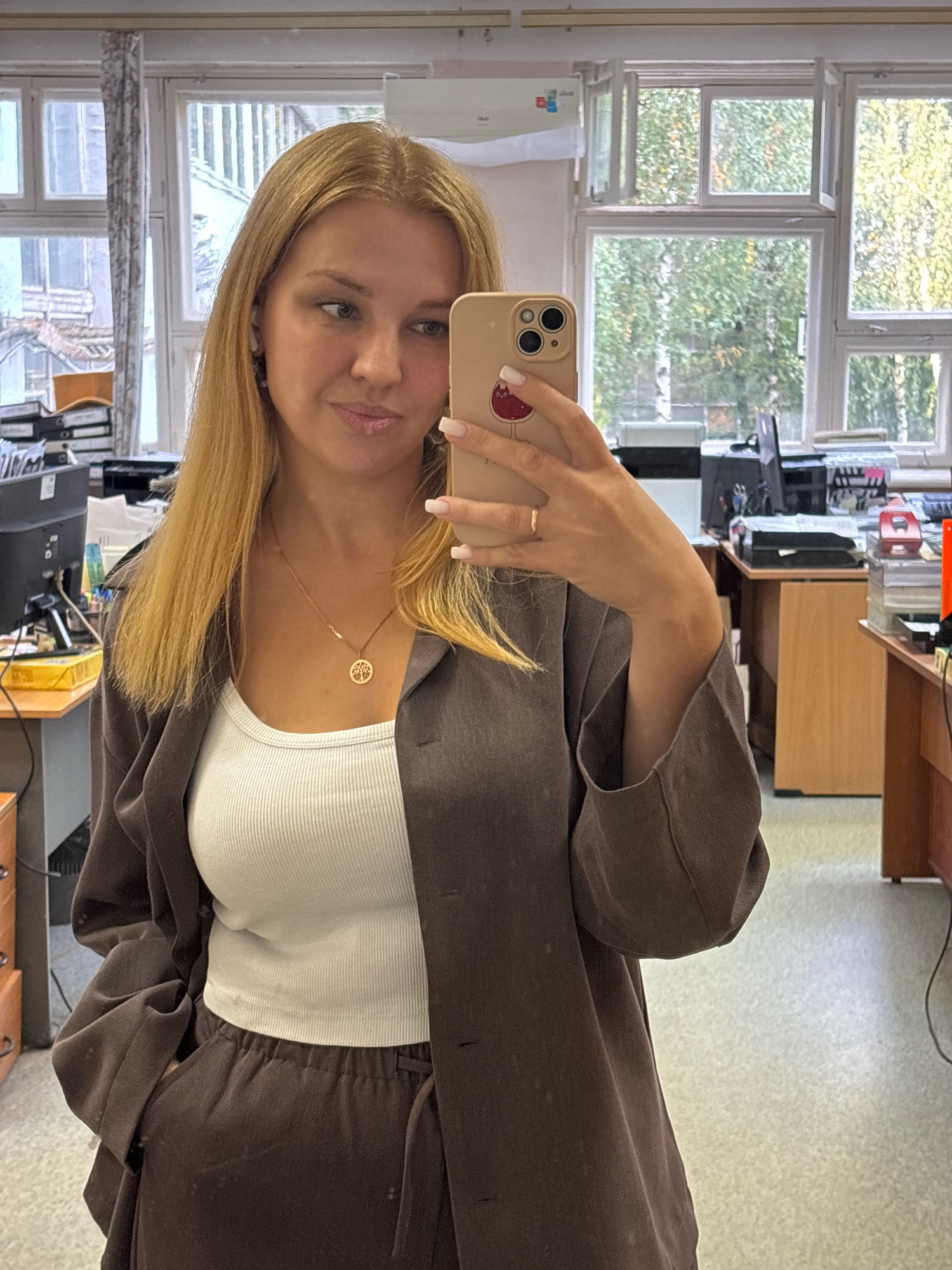 A woman is taking a selfie in an office environment. She has blonde hair and is wearing a white ribbed tank top, a brown blazer, and brown pants. She is also wearing a necklace with a circular pendant featuring a tree design. Her left hand is in her pocket, and her right hand is holding a smartphone. In the background, there are windows with trees visible outside, office desks with computers and printers, and filing cabinets. The location is identified as Rusanovo, Russia.