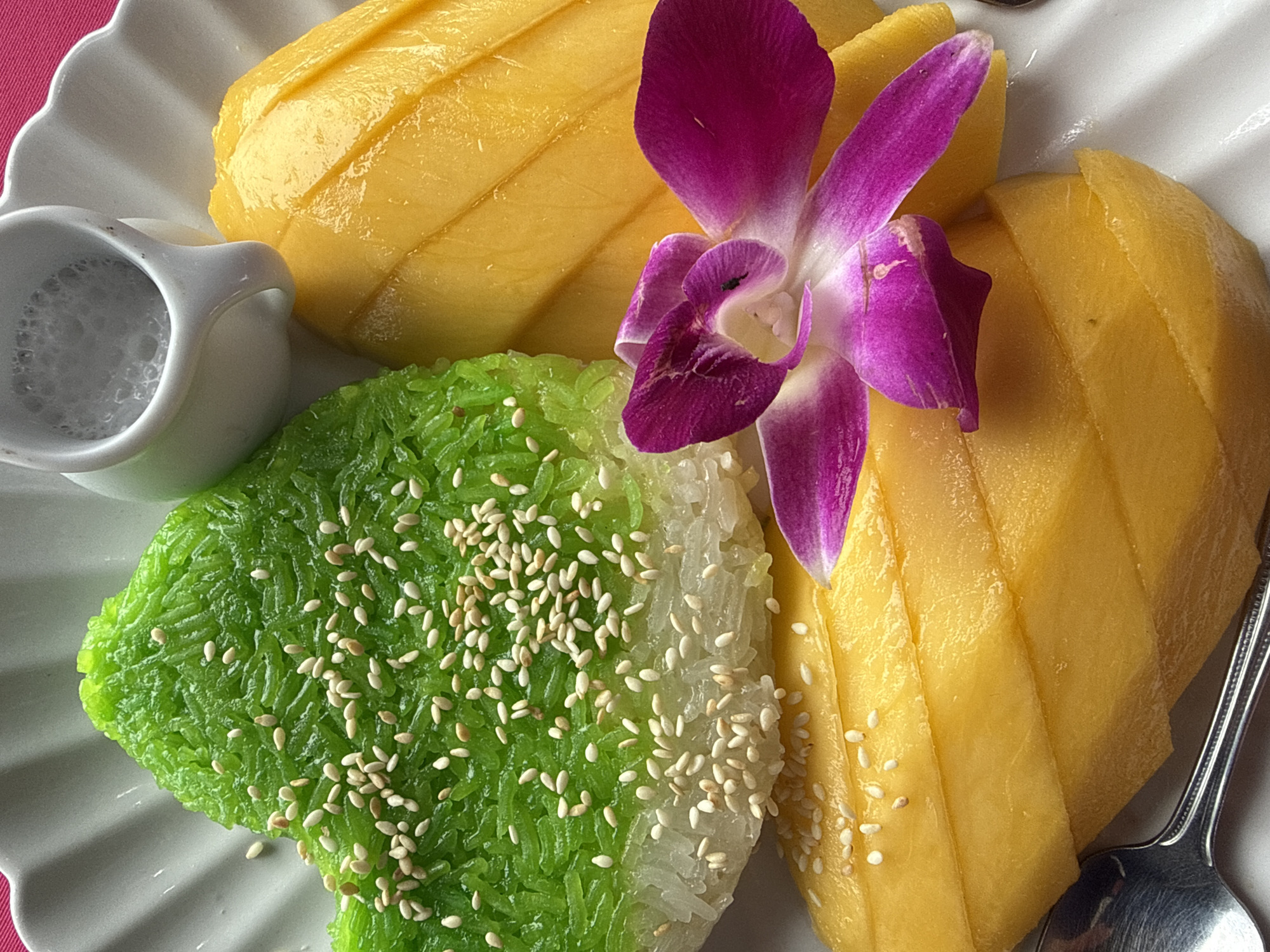 A close-up, overhead shot showcases a plate of mango sticky rice, a popular Thai dessert, presented on a white, fluted plate. The vibrant green sticky rice, molded into a heart shape, is topped with a generous scattering of toasted sesame seeds. To the right of the rice, two generous slices of ripe yellow mango are arranged. A single, deep purple orchid with white accents rests delicately on top of the mango slices, adding a touch of elegance to the presentation. A small, white ceramic pitcher, containing a white liquid, possibly coconut milk, is positioned in the upper right corner of the plate. A silver spoon is partially visible in the lower left corner. The image was captured in Karon, Thailand.