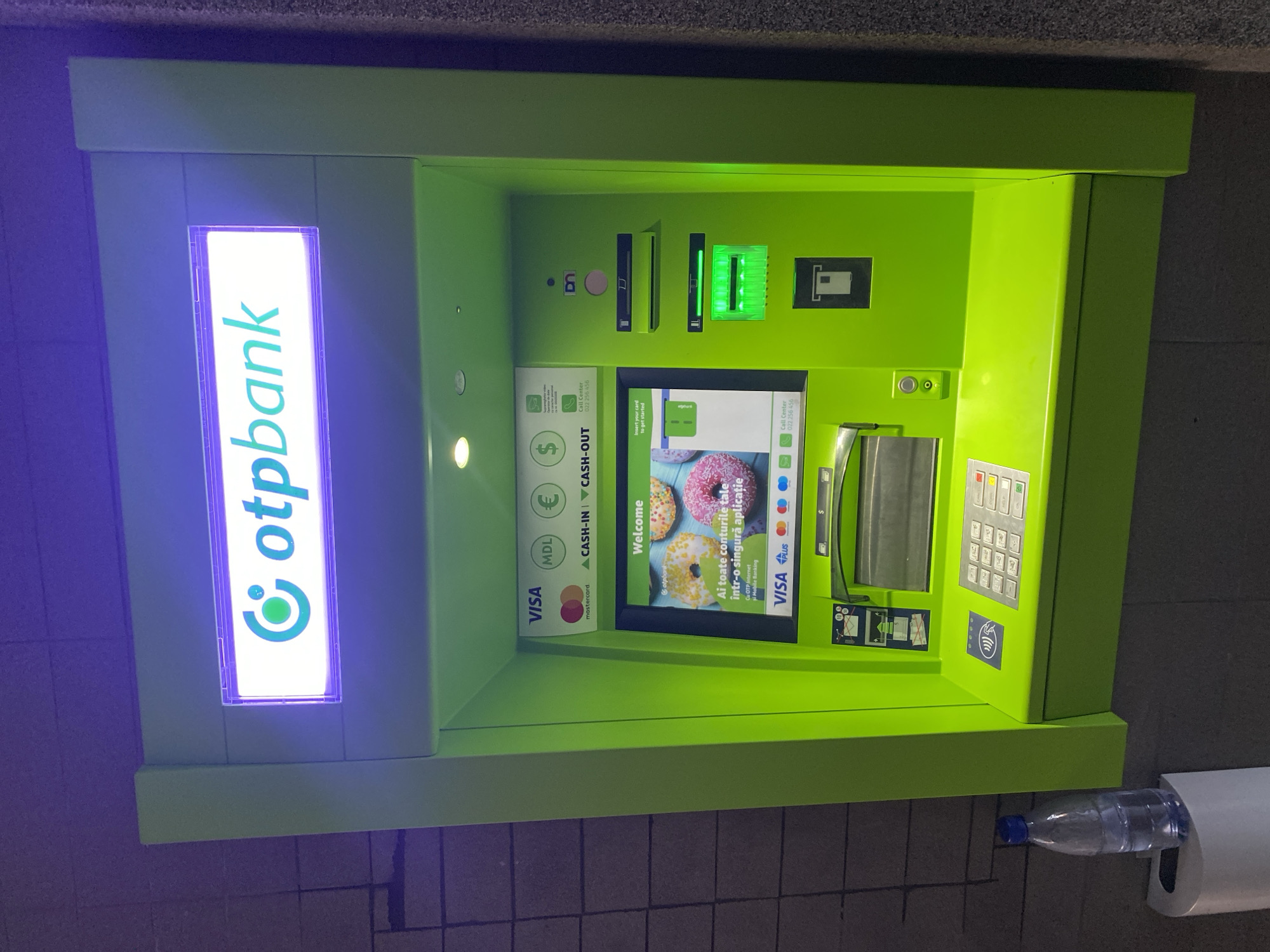 The image shows an OTP Bank ATM in Chișinău, Moldova. The ATM is bright green and has the OTP Bank logo illuminated at the top. The screen displays a welcome message in Romanian and an image of donuts. Below the screen are card slots, a keypad, and a cash dispenser. A bottle of water is placed on a roll of paper towels to the left of the ATM. The ATM is situated in an indoor or covered outdoor area with a textured wall in the background. The lighting suggests it is nighttime or dimly lit.
