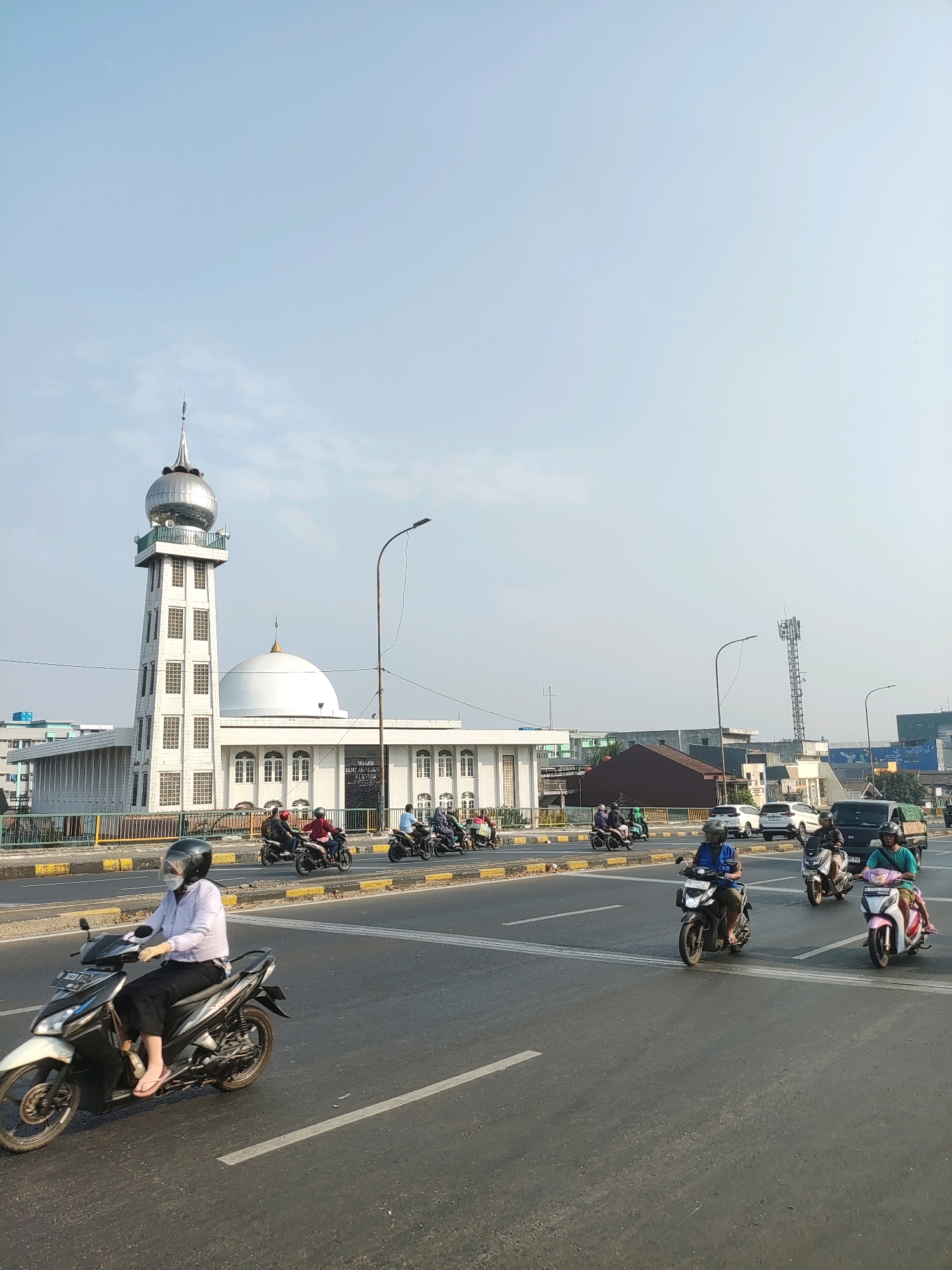 The image shows a street scene in East Jakarta, Indonesia, with a prominent mosque in the background. The mosque is a white building with a large dome and a tall, slender minaret topped with a silver finial. A street runs in front of the mosque, with several lanes of traffic. Numerous motorcycles are visible, with riders wearing helmets and masks, suggesting a concern for safety and possibly public health. Cars and a truck are also present in the traffic. The sky is clear and blue, indicating a sunny day. The overall scene depicts a typical urban environment in Indonesia, characterized by religious architecture and active street life.