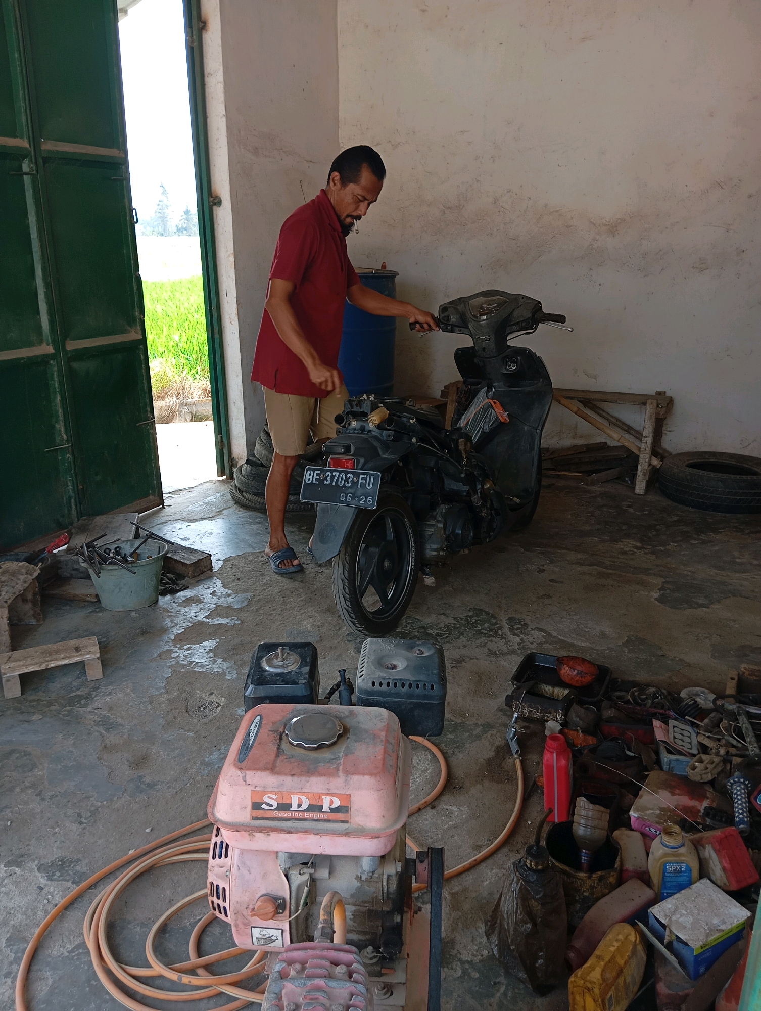 In a workshop setting in Leman Benawi, Indonesia, a man in a red polo shirt and tan shorts is working on a black motorcycle. The workshop appears to be an open-air structure with a concrete floor, partially covered by a roof. To the left, a large green door stands ajar, revealing a bright, sunlit field with green crops and trees in the distance.
The man, with short dark hair and a mustache, is holding onto the handlebars of the motorcycle, which is partially disassembled. Various tools and parts are scattered around the workspace. In the foreground, a pink gasoline engine with the "SDP Gasoline Engine" logo sits next to a coiled orange hose. Other objects in the workshop include stacked tires, a blue barrel, a bucket filled with metal rods, wooden planks, and an assortment of containers and tools, some of which appear to be used for vehicle maintenance or repair. The overall scene depicts a functional, albeit somewhat cluttered, repair area.