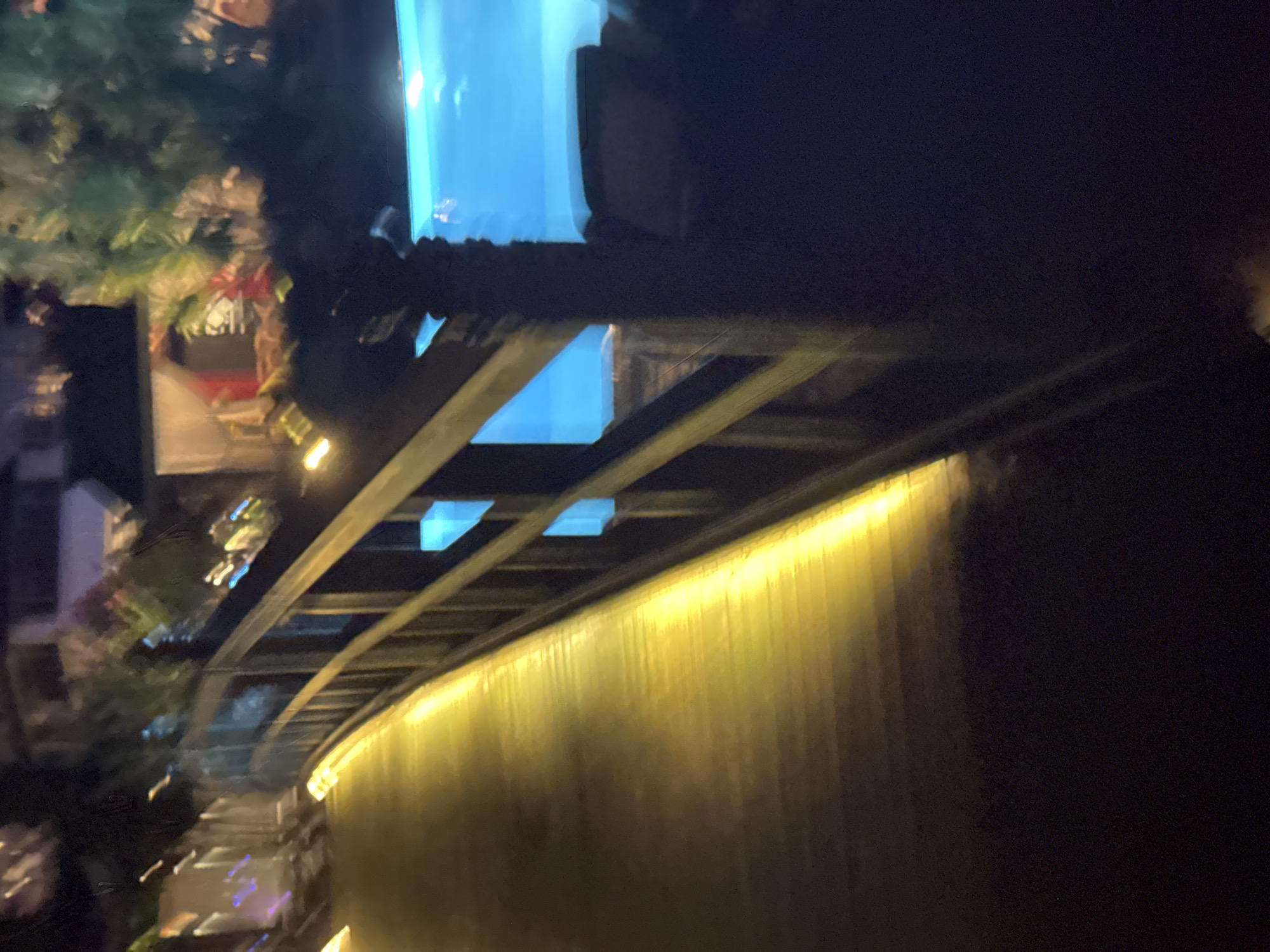 A nighttime shot shows a wooden bridge with yellow lighting along its edge. To the right of the bridge, a bright blue swimming pool is visible. In the background, there are blurred structures and lush greenery. The image is taken at night and appears to be in Aksu, Turkey, based on the provided context.