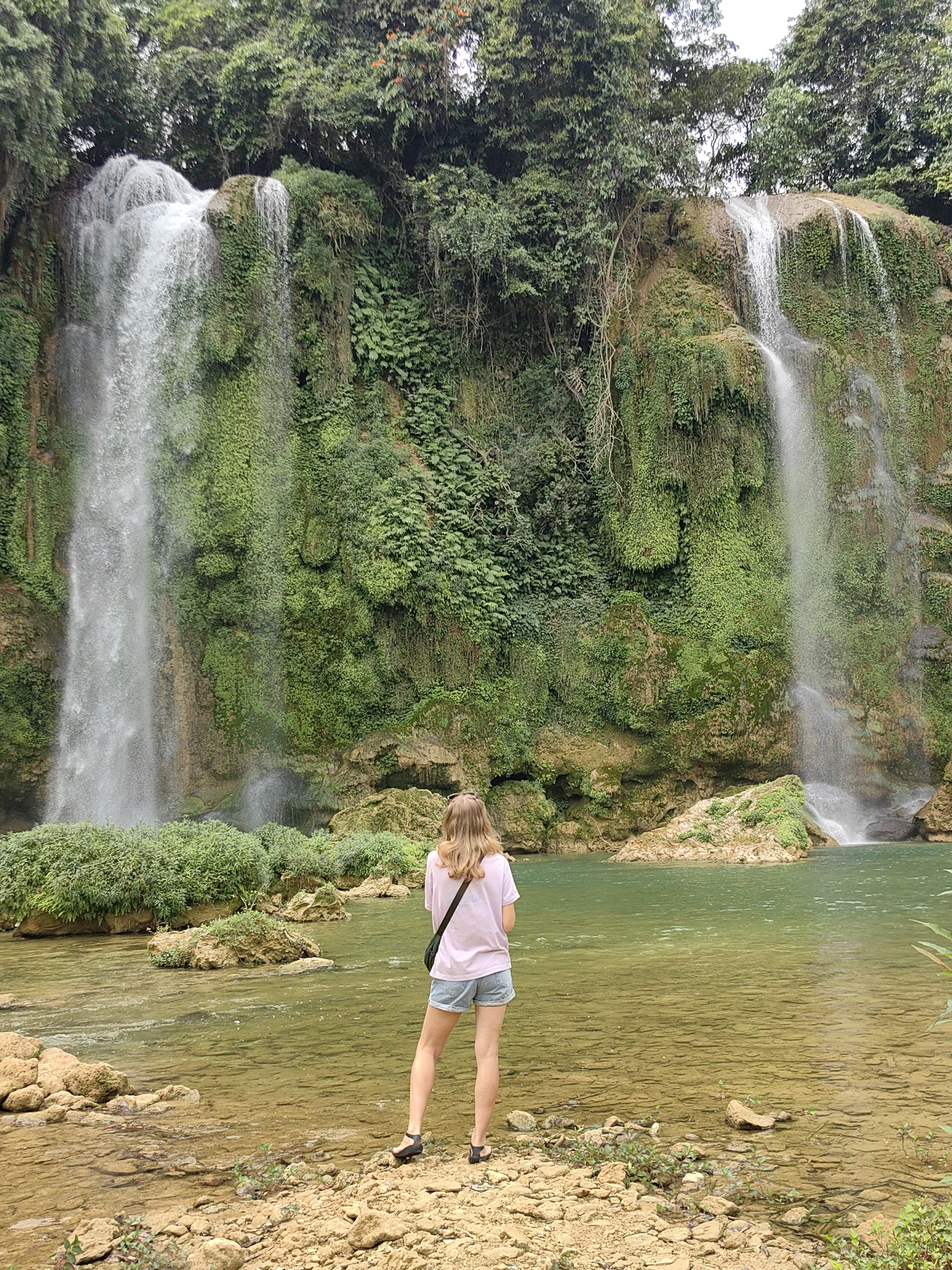A woman stands in shallow, clear water, viewed from behind, facing a wide waterfall in Daxin County, China. The waterfall is composed of two main streams, one on the left being significantly wider than the one on the right. The rock faces behind the waterfalls are covered in dense green vegetation. The water in the foreground appears calm and clear, with small rocks and plants along the edge where the woman is standing. She is wearing a light-colored t-shirt, denim shorts, and dark sandals. A black bag strap is visible across her back. The scene is set in a lush, green environment.