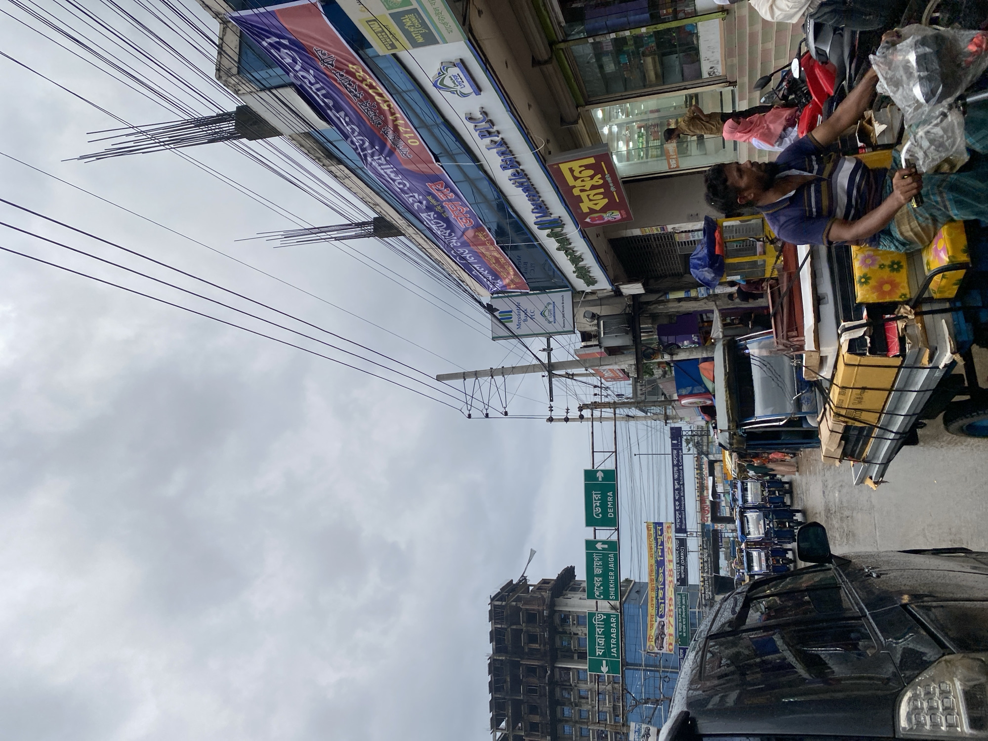 A street scene in Dhaka, Bangladesh, with a cloudy sky. The image shows a busy road with various modes of transportation, including rickshaws, a car, and a motorcycle. In the foreground, a man is driving a rickshaw laden with boxes. In the background, buildings line the street, some of which are under construction. Traffic signs indicating directions to "Jatrabori," "Shekher Jaiga," and "Demra" are visible. A banner on one of the buildings advertises "Mercantile Bank PLC." The overall impression is one of a bustling urban environment with ongoing development and daily life.