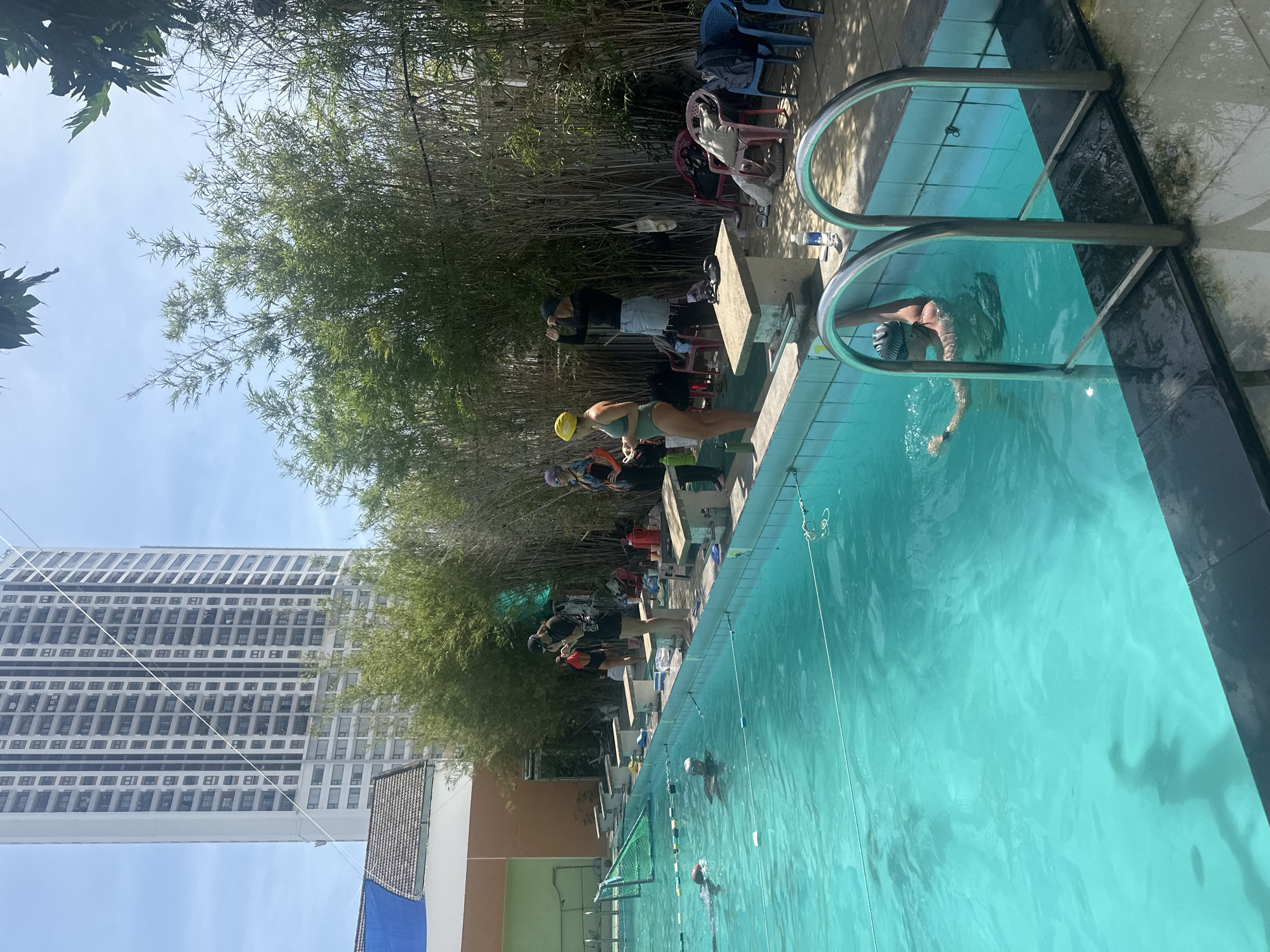 The image depicts an outdoor swimming pool area with several individuals, likely engaged in a swimming activity or lesson. A tall, modern residential building dominates the background, suggesting an urban or semi-urban setting. The pool itself is filled with bright turquoise water, and swim lanes are marked by ropes and buoys. Around the pool, there are several diving blocks and benches, as well as some chairs. Lush green bamboo plants line the back of the pool area, providing a natural screen. One person is visible in the water, swimming towards the foreground. Several other individuals are standing on the pool deck, some in swimwear, appearing to be waiting their turn or observing. The overall atmosphere suggests a sunny day at a public or private swimming facility.