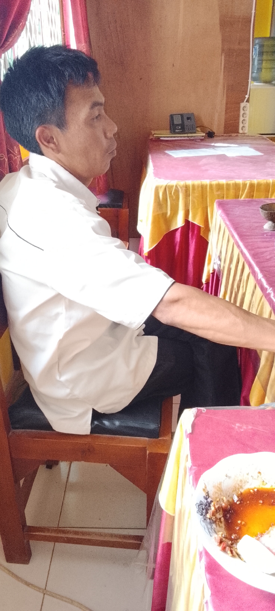 A man sits on a wooden chair with a black cushion, viewed from the side. He is wearing a white short-sleeved collared shirt and black pants. His hair is dark and short. He is seated at a table covered with a magenta tablecloth, on which a plate with food and sauce is visible. In the background, another table is visible, also covered with a magenta and yellow tablecloth. On this table, a landline telephone rests on a stack of papers. The room appears to be indoors, with a wooden wall and possibly a window with red curtains on the left. The floor is tiled. The scene is likely in a community space or a simple dining area. Without further context, the specific location in Jepara, Indonesia cannot be determined.