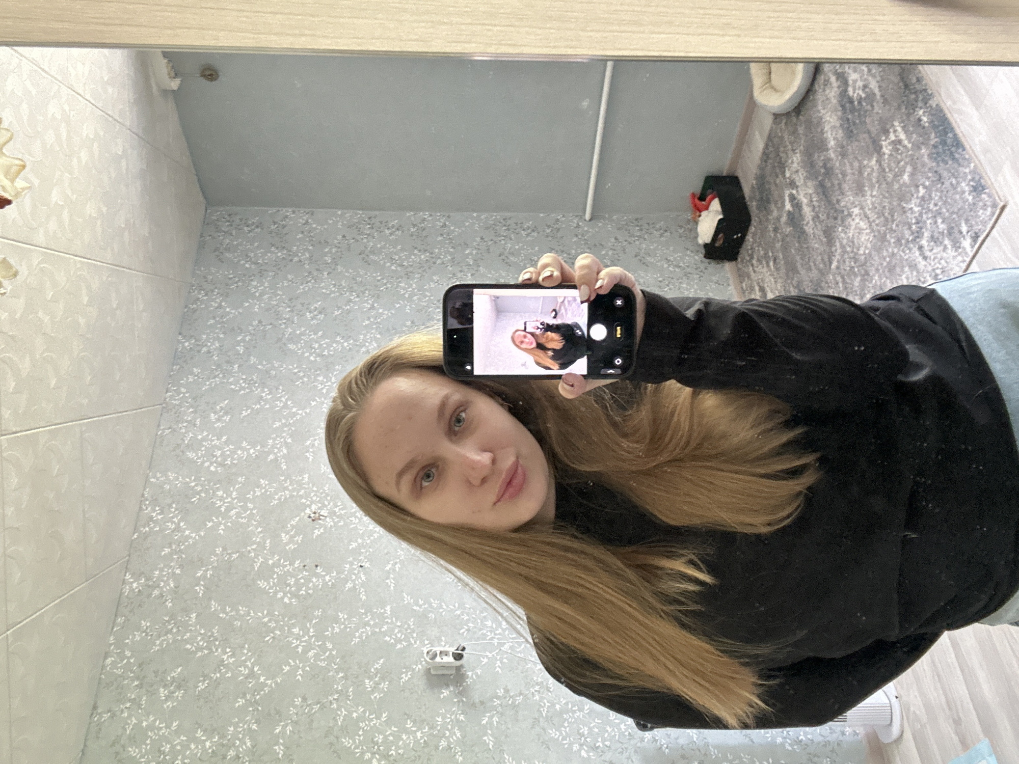The image depicts a woman taking a selfie in a mirror. She is holding her phone up, displaying a smaller image of herself on the screen. The woman has blonde hair and is wearing a black sweater.
The reflection in the mirror reveals a bathroom setting. The walls are covered in a light blue wallpaper with a white floral pattern. To the left, there are white tiled walls. In the background, on the floor, there is a gray rug, a pet bed, and a small black container with red and white items inside. A white baseboard heater is visible in the bottom right corner. The location is in Chaikovsky, Russia.
