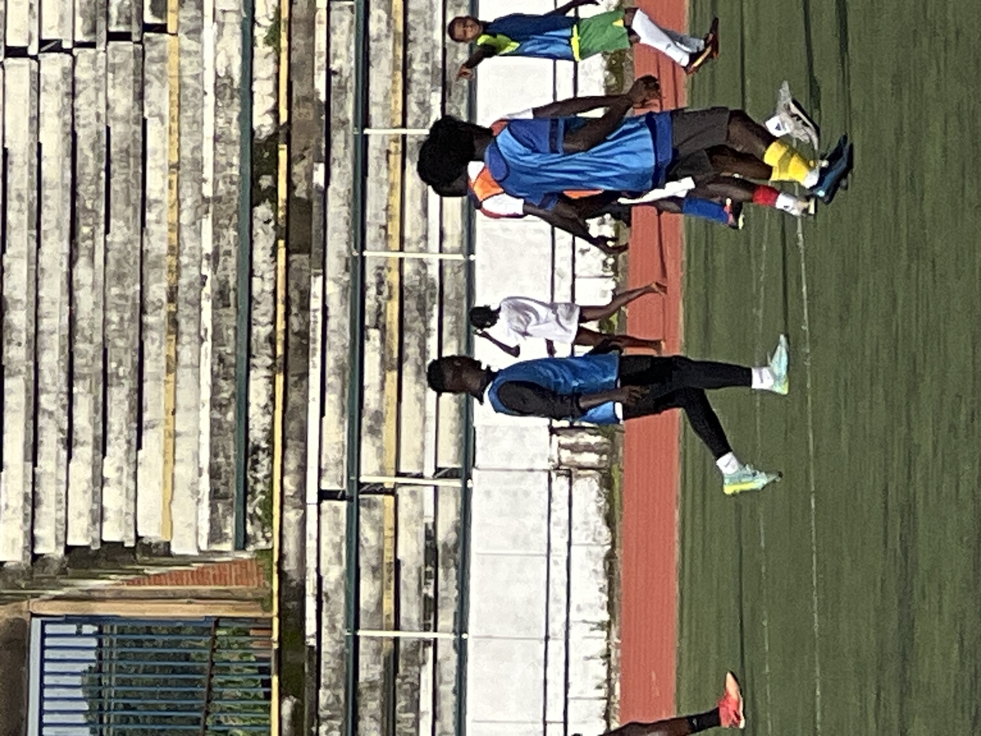 In New Stadium, Nigeria, several individuals are on a grassy sports field, possibly a soccer field, with a red track running alongside it. In the background, there are tiered concrete bleachers with some overgrown vegetation. A fence with blue bars is visible to the left of the bleachers. Several people, appearing to be young men and women, are scattered across the field, some wearing athletic attire. One person in the foreground on the right is wearing a blue vest over a black shirt, dark shorts, yellow socks, and dark athletic shoes, and is looking to their left. Another individual in the foreground on the left is wearing a blue vest over a black shirt, black pants, and light blue athletic shoes. In the middle ground, a person in a white shirt and shorts is walking away from the camera. Another person in a blue vest is partially visible behind them. To the far right, a person in a blue and green jersey with green shorts and white socks is pointing to their left. The scene suggests a sports practice or gathering on a sunny day.