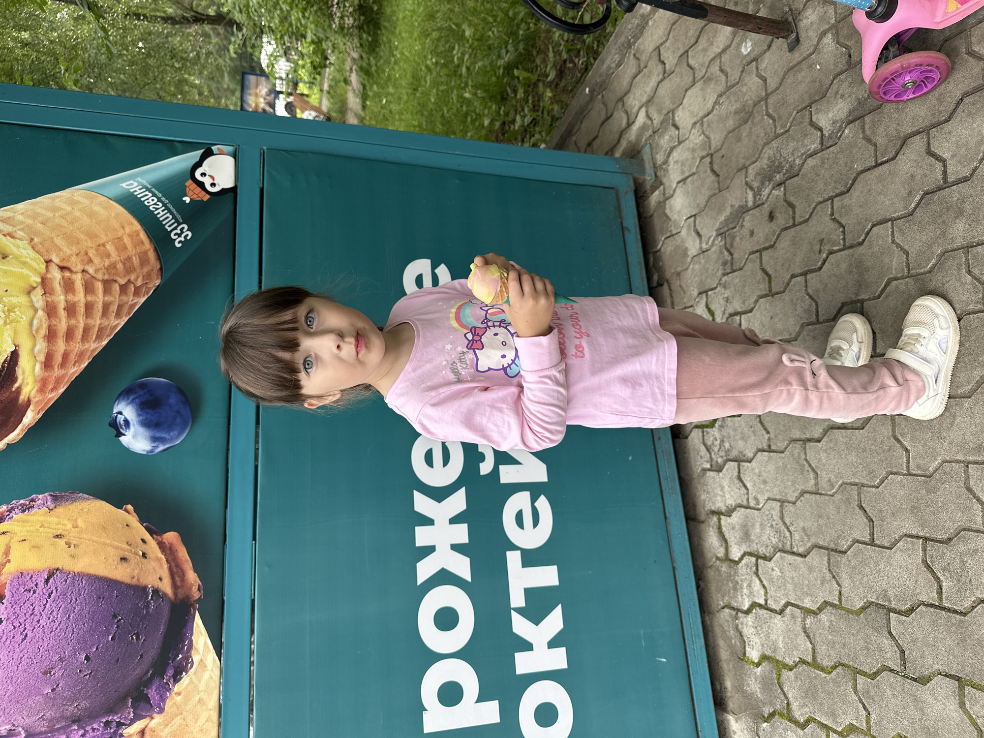 In Chaikovsky, Russia, a young girl with blue eyes and dark hair stands in front of a teal-colored advertisement featuring large ice cream cones and a blueberry. She is wearing a pink long-sleeved shirt with a Hello Kitty graphic and pink pants, and she holds a small ice cream cone in her right hand. White-patterned pavement is visible beneath her feet, and a pink scooter is partially visible to the right of the frame. Trees and greenery are present in the background.