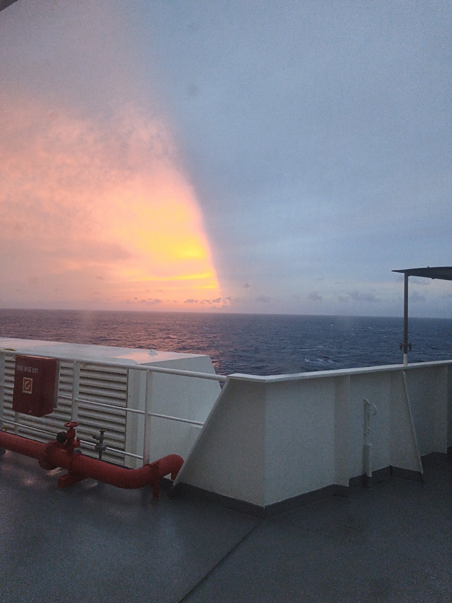 The image shows a sunset over the ocean, viewed from the deck of a ship. The sky is illuminated with warm orange and yellow hues on the left, transitioning to a pale blue on the right. The sea is a deep blue, with gentle waves. In the foreground, there is a white railing and deck. A red fire hose and a red box labeled "FIRE HOSE BOX" are visible on the deck. The scene is serene and atmospheric, suggesting a moment of quiet reflection. The image does not depict the city of Tongyeong-si, South Korea.