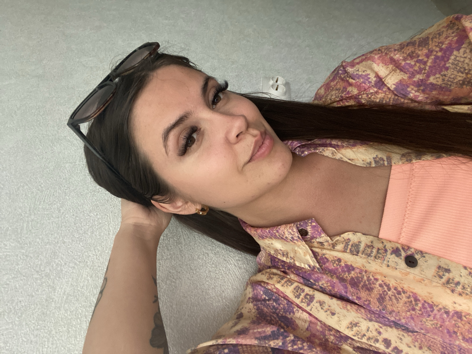A young woman with long, dark hair is shown from the chest up, leaning her head back against a light-colored, textured wall. She is wearing a colorful, patterned button-up shirt over a peach-colored top. Her left arm is bent, with her hand resting behind her head, revealing a tattoo on her upper arm. Sunglasses are perched on top of her head. A gold hoop earring is visible on her right ear. Her gaze is directed slightly off-camera, and she has a subtle smile. A white electrical outlet is visible on the wall behind her to the right. The overall lighting suggests an indoor environment.