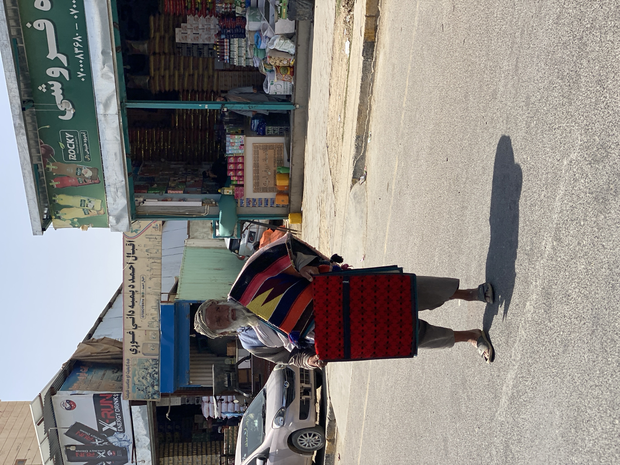 An elderly Afghan man, dressed in a traditional turban and gray tunic, stands in the middle of a paved street in Kandahar, Afghanistan. He is holding a vibrantly colored, patterned textile draped over his left arm and a red, intricately patterned fabric with geometric designs in front of him, likely for sale. The man has a long, white beard and appears to be looking towards the viewer with a neutral expression.
Behind him, a storefront displays various goods, including what appears to be sacks of grain or rice stacked high. Several bottles of what may be beverages are visible on a counter inside the store. A beige car is parked on the side of the road to the left of the man, and a street curb with a dirty sidewalk is visible in the foreground. The scene is illuminated by bright sunlight, casting a distinct shadow of the man on the asphalt. The architecture suggests a commercial area in an urban setting.