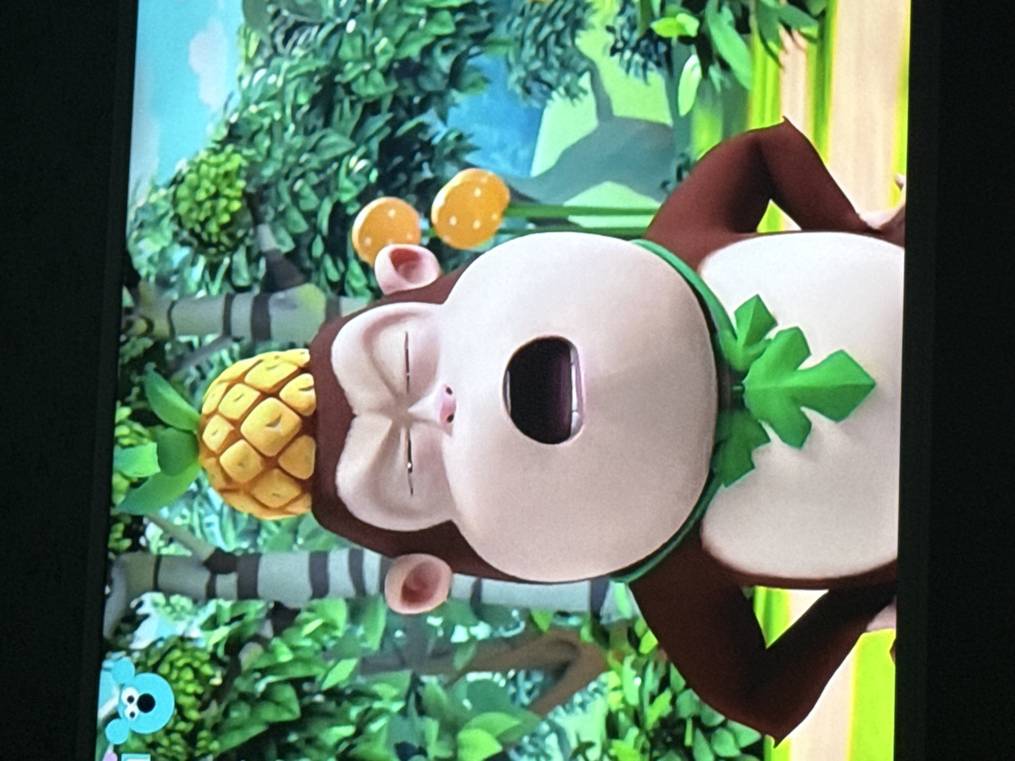 The image depicts a cartoon monkey character with a pineapple on its head. The monkey has brown fur around its face and arms, a white face and chest, and is wearing a green leafy collar. Two yellow fruits with polka dots are visible near the monkey's head. The background consists of lush green foliage, possibly representing a jungle or forest setting, with some light blue sky visible. In the bottom left corner is a blue cartoon character icon. The image appears to be a still frame from a cartoon or animated show. There is no indication of the location of Mytishchi, Russia.