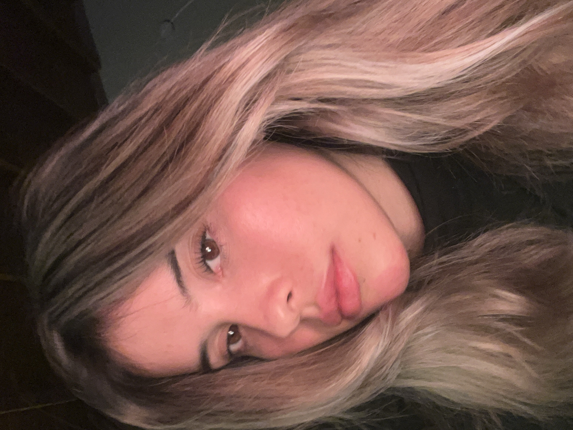 A close-up, medium shot captures a young woman's face, angled slightly to her left, against a dark, out-of-focus background. She has long, wavy, blonde hair with lighter streaks, some of which fall across her face. Her skin has a warm, pinkish tone, possibly due to lighting. She is wearing a dark, high-necked top. Her eyes are dark brown and appear to be looking slightly downward. Her eyebrows are dark and well-defined, and she has long eyelashes. Her lips are full and appear to be slightly parted. The lighting casts a warm glow on her face, with a pinkish hue visible on her left cheek and forehead. The image's composition focuses on her facial features, particularly her eyes, nose, and mouth. There is no identifiable location in Sumalao, Argentina.