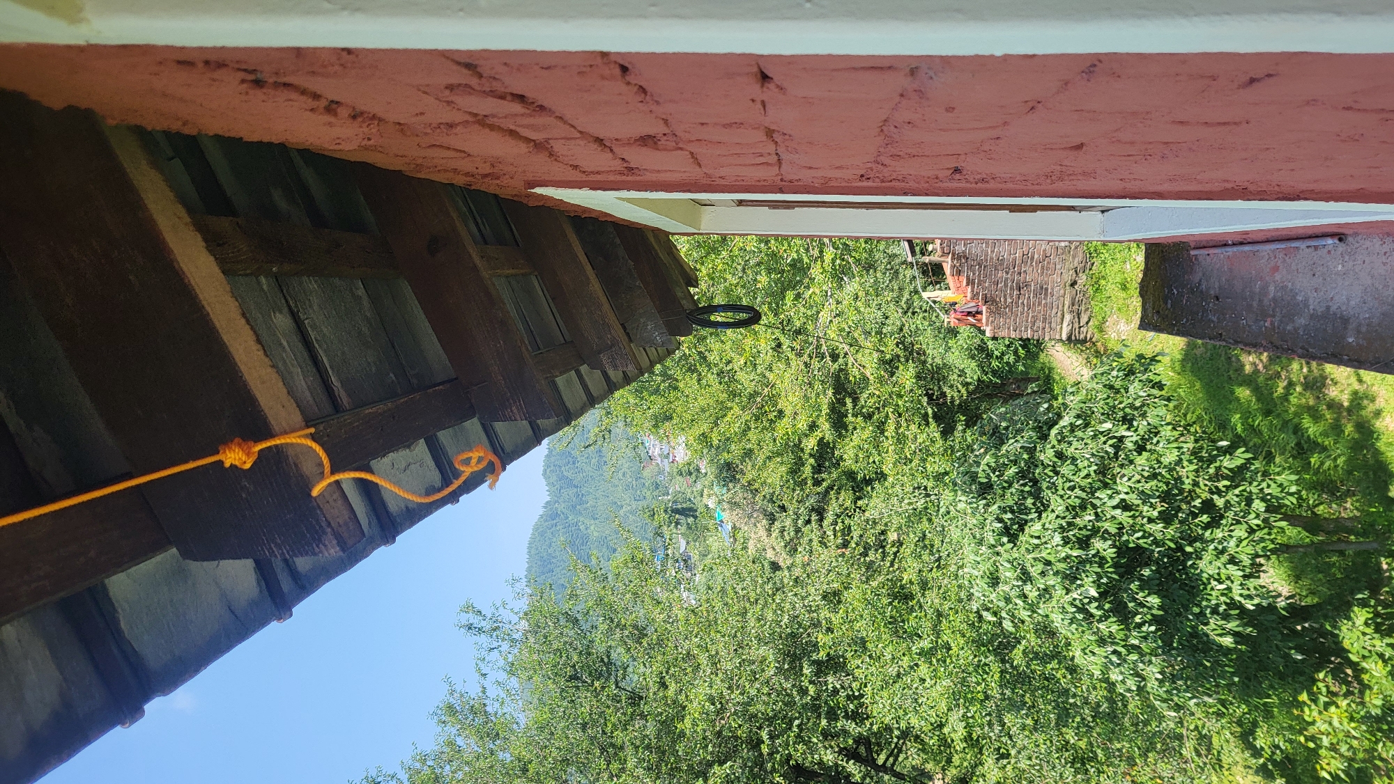 From an elevated perspective, the photograph captures a segment of a building's structure, presumably in Haini, India, as suggested by the visual cues in the distant landscape. The scene includes a section of a wooden roof, an orange rope tied to its beams with knot details, and a red-brick exterior wall. A black ring hangs from a beam. The background reveals dense green trees and vegetation, suggesting a rural or mountainous setting. In the distance, there's a glimpse of a small building. The sky is partially visible, displaying a clear blue.