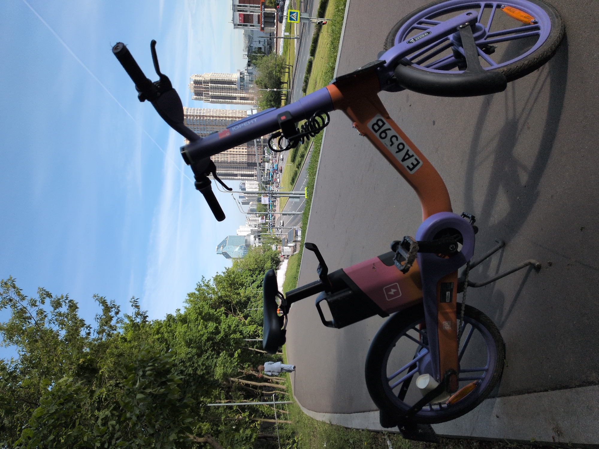 A photograph taken in Moscow, Russia, shows a V-shaped electric scooter on a paved path. The scooter has an orange stem with the code “EA659B” printed on it. The tires and other parts are lavender or purple. Trees line the left side of the path, and buildings are visible in the distance.