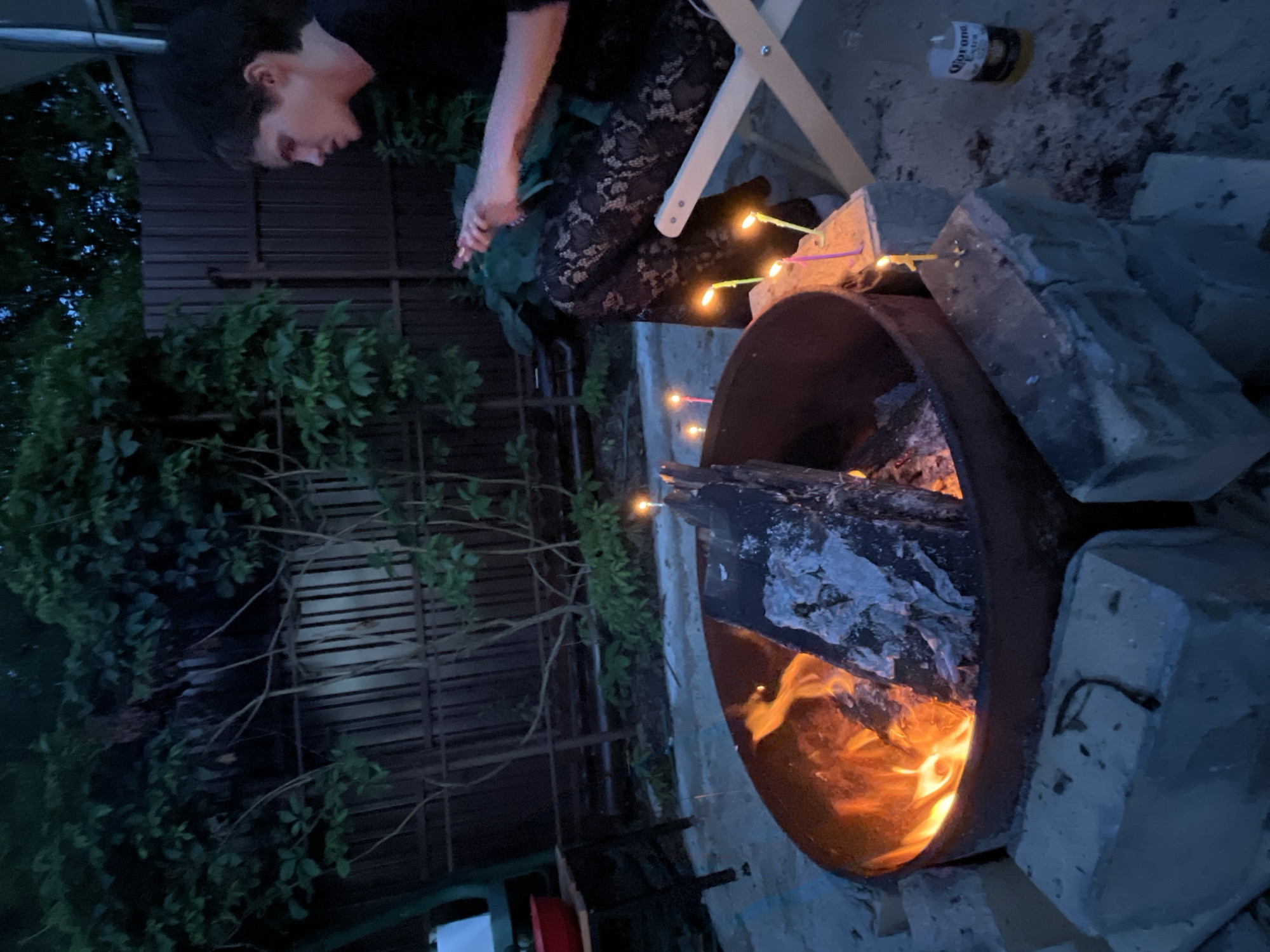In the dimly lit outdoor setting of Вельяминово, Russia, a young person with dark hair sits on a folding chair, facing away from the camera, towards a bonfire. They are wearing a black top and patterned black lace pants. The bonfire, contained within a metal ring on a bed of sand and surrounded by concrete blocks, burns with bright orange flames and emits smoke. Several lit candles of varying colors are placed on the concrete blocks near the fire. To the right of the scene, a can of Corona Extra is visible on the ground. The background features a dark wooden fence and lush green foliage.